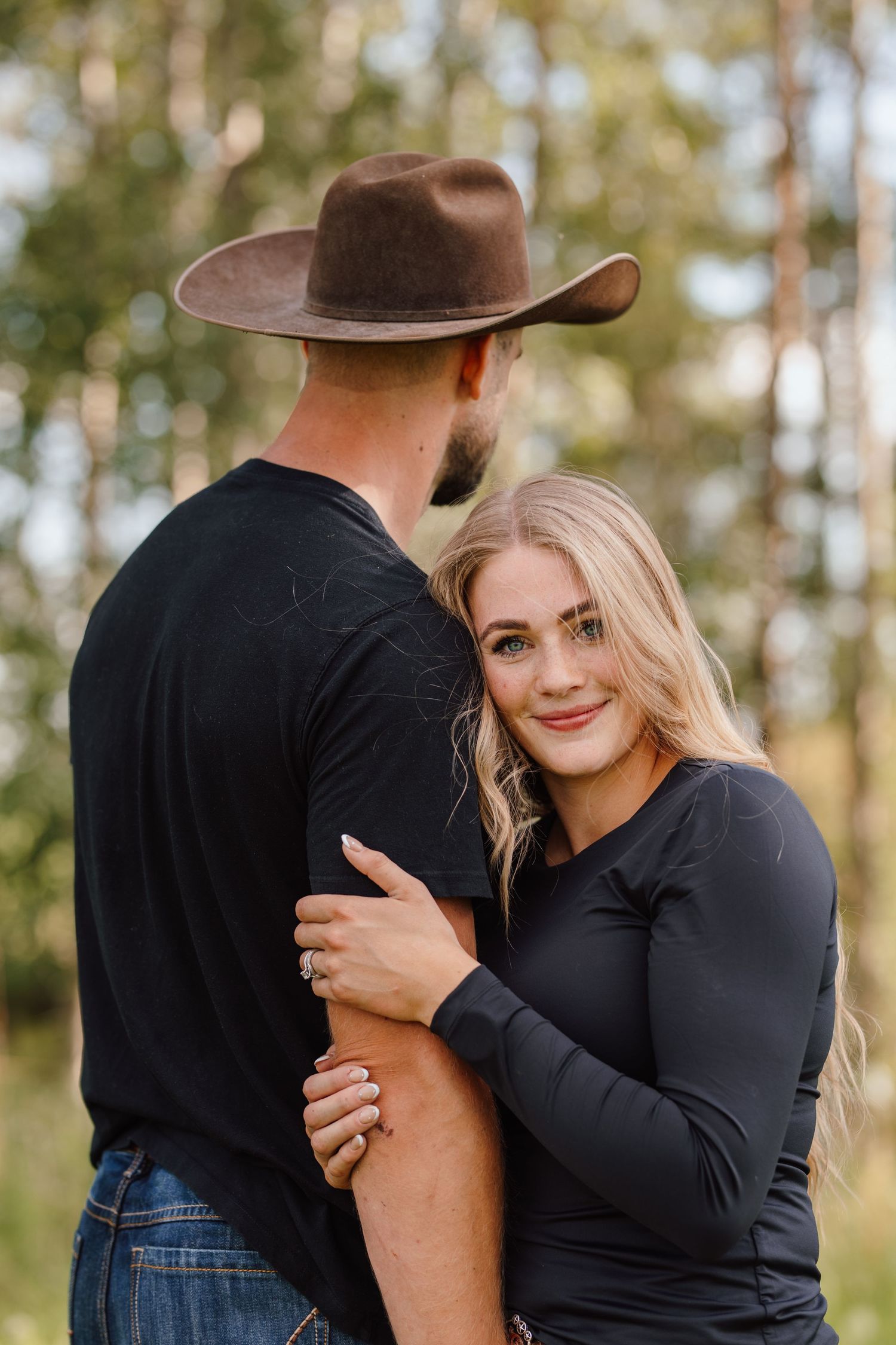 A blonde woman in a black top embraces a man in a brown cowboy hat and black shirt outdoors among trees.