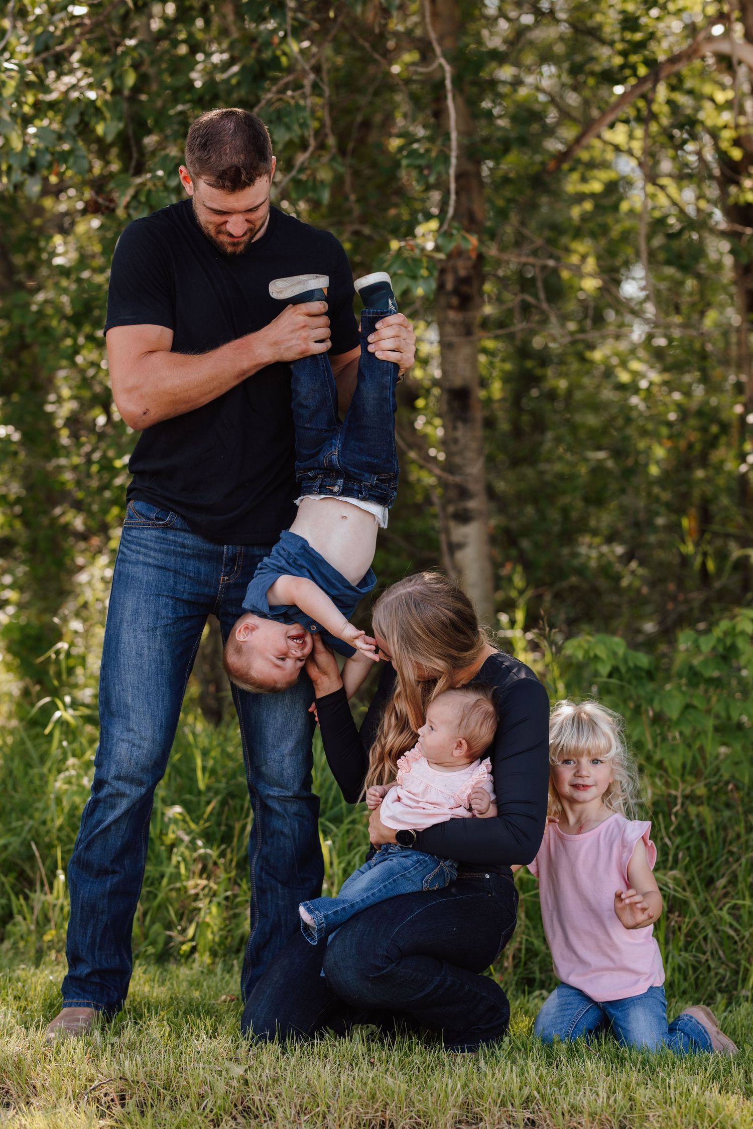 Family of five enjoying outdoor fun, dad playfully holding child upside down while mom kneels with baby and young daughter smiles.