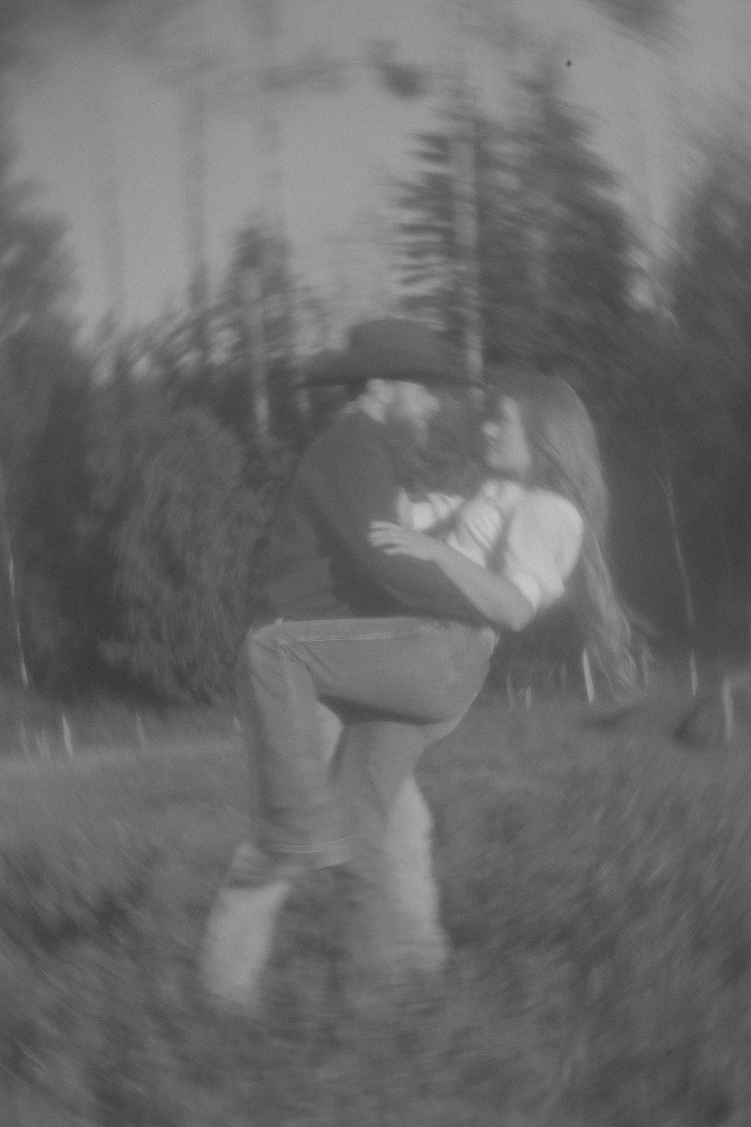 Black and white photo of a couple embracing and kissing outdoors among trees in a dreamy blurred romantic scene.