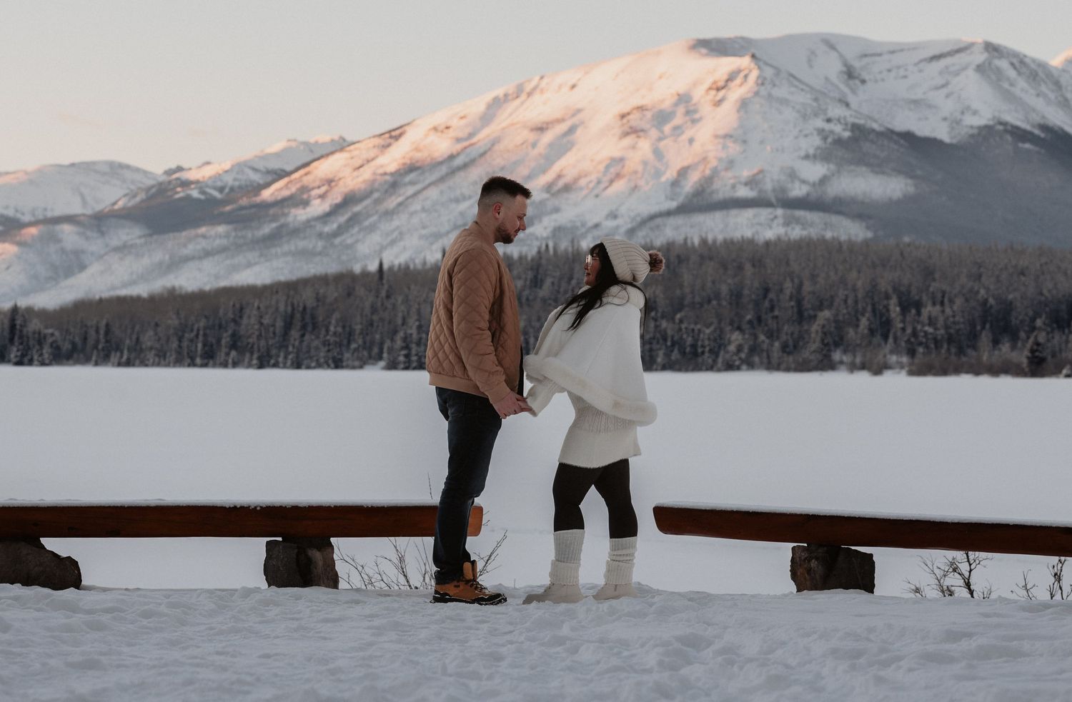 Couple holding hands in snowy mountain landscape at sunrise, standing near a wooden fence with snow-capped peaks behind them.