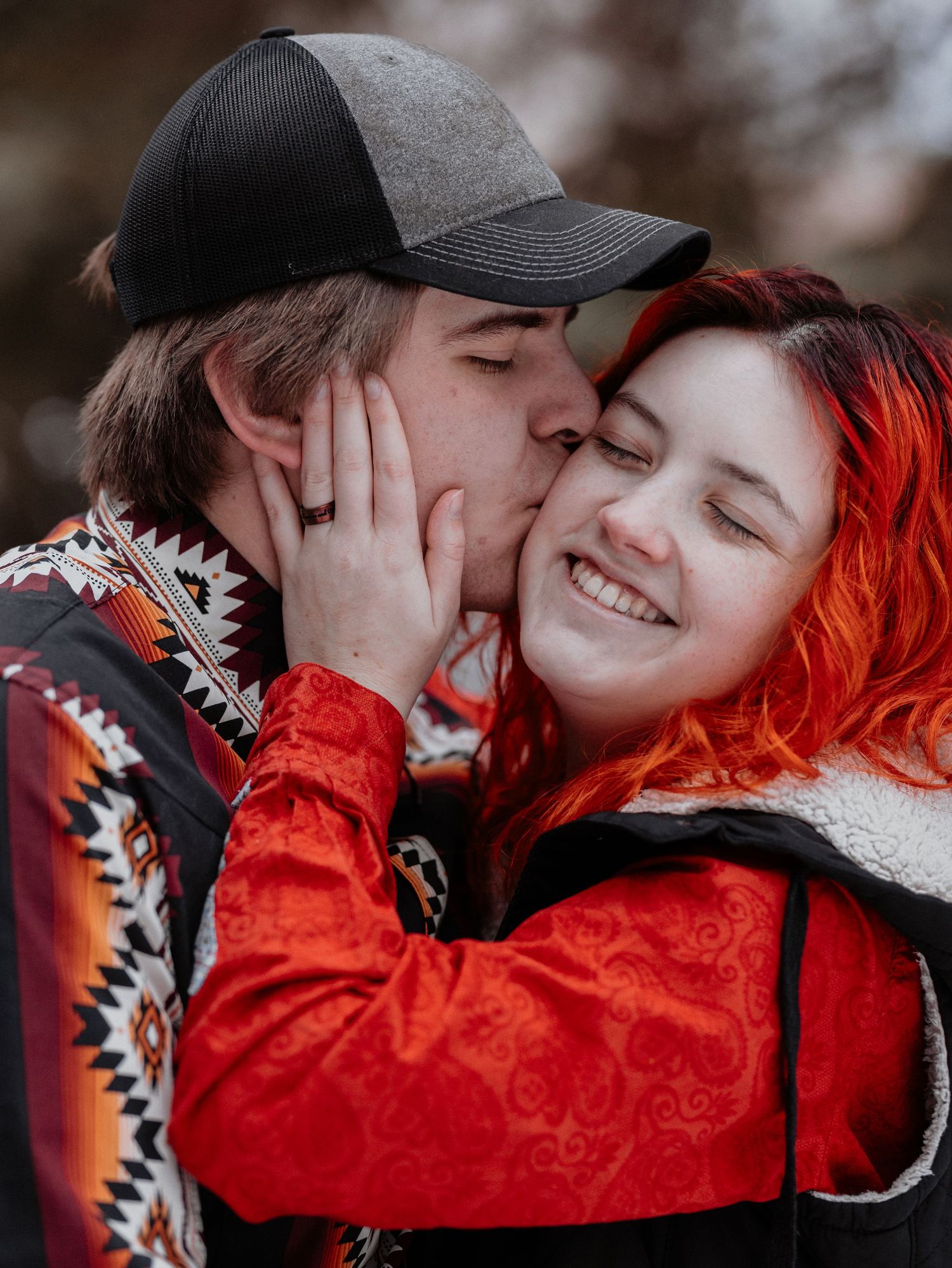 Man kissing smiling woman with vibrant red-orange hair in cozy winter outdoor portrait.
