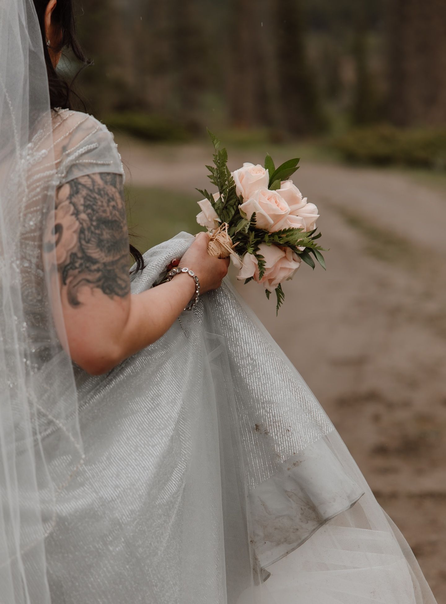 Tattooed bride holding delicate pink rose bouquet under flowing veil in forest setting.