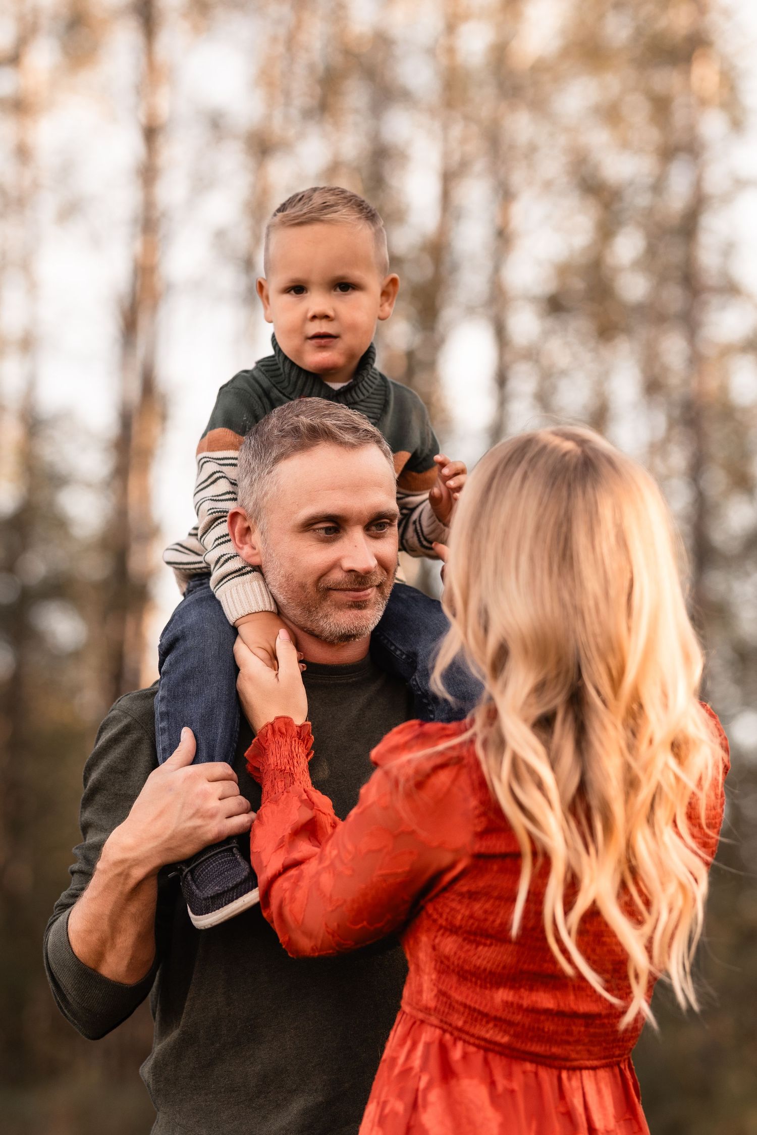 A happy family outdoors in autumn, father carrying toddler on shoulders while blonde mother in red dress looks on.
