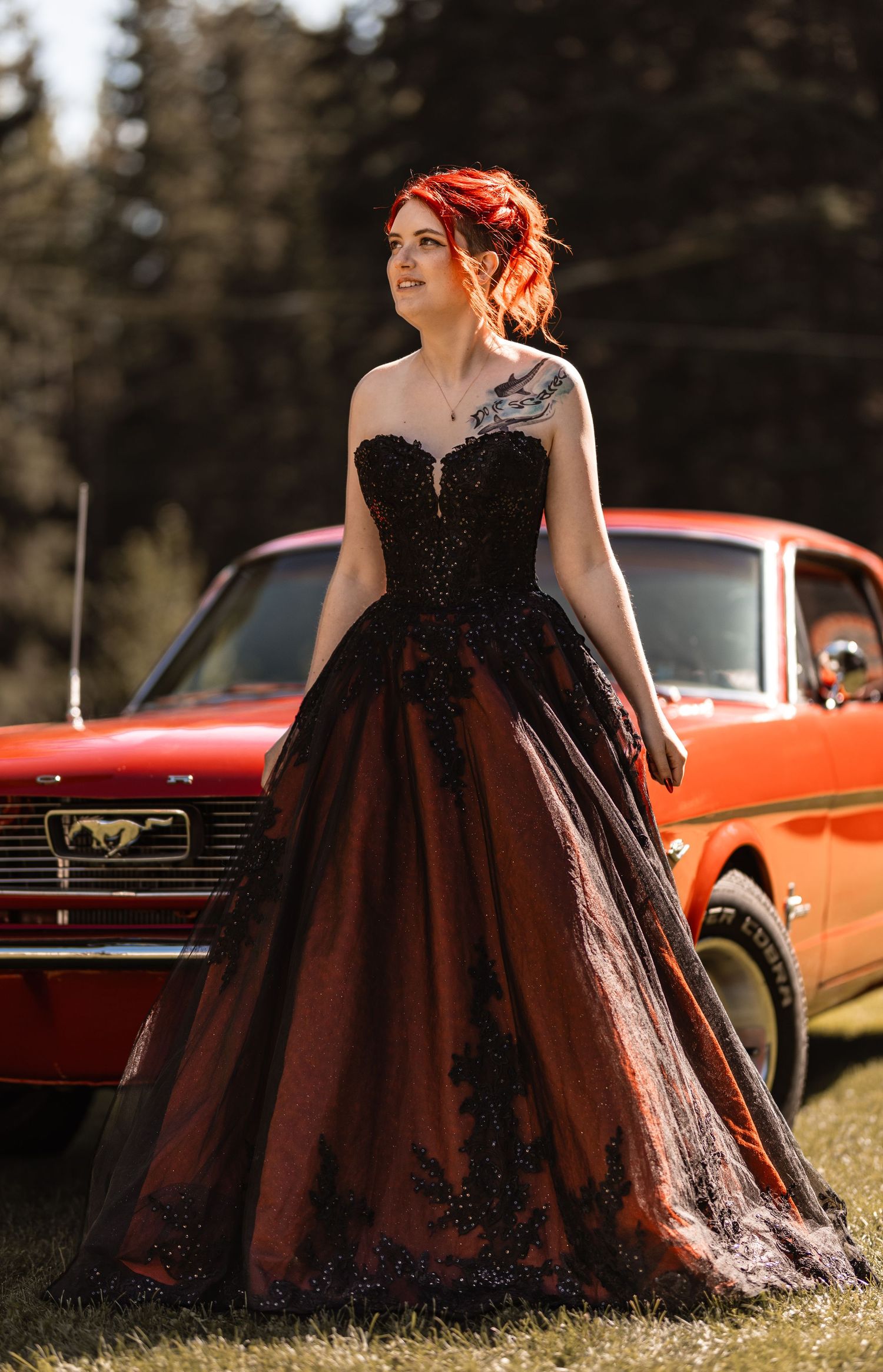 Red-haired woman in black lace ballgown poses beside vintage red Ford Mustang outdoors.