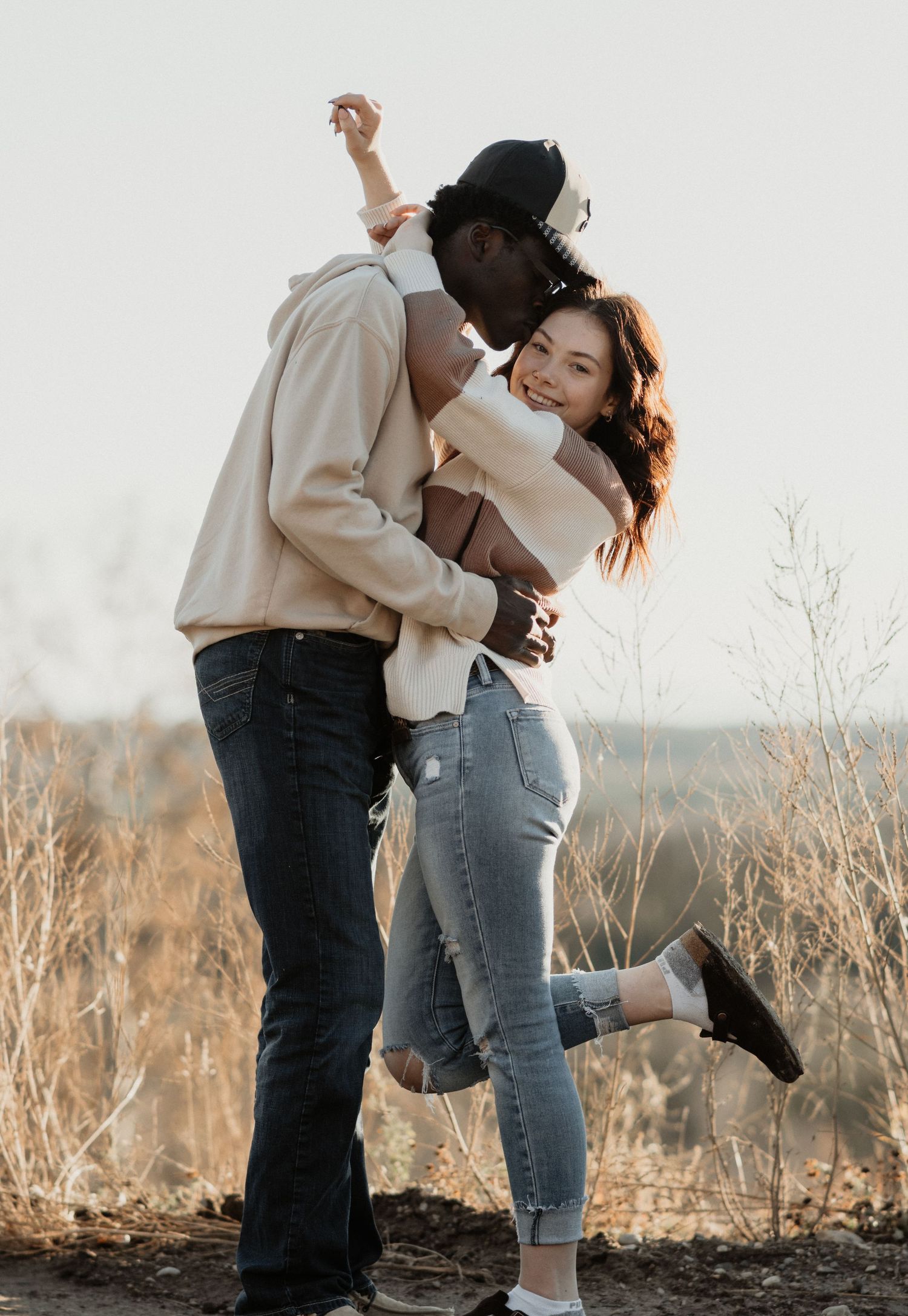Interracial couple embracing and dancing outdoors in a golden field during sunset engagement photo session.
