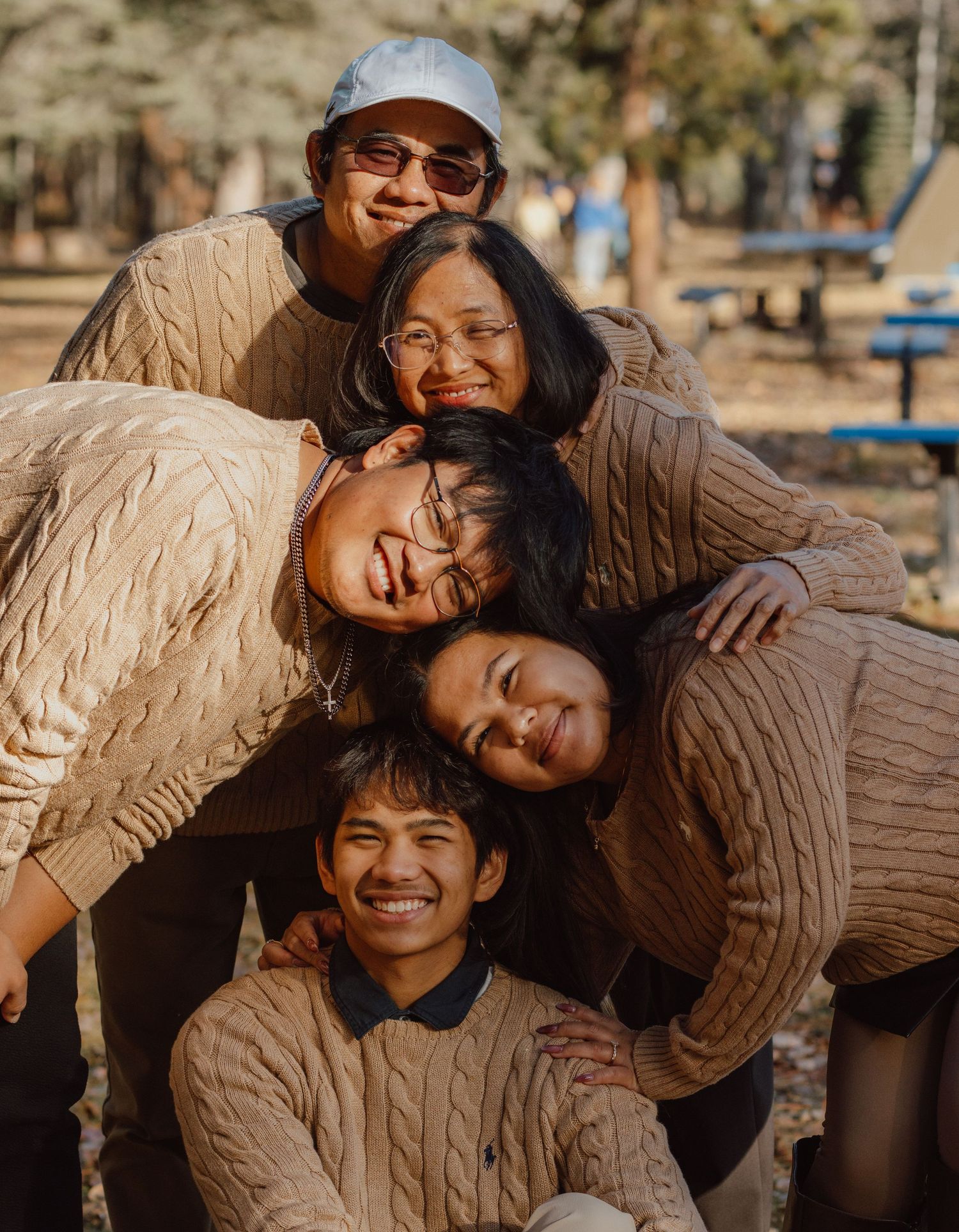 Five friends in matching beige cable-knit sweaters huddle together smiling in a sunny outdoor park setting.