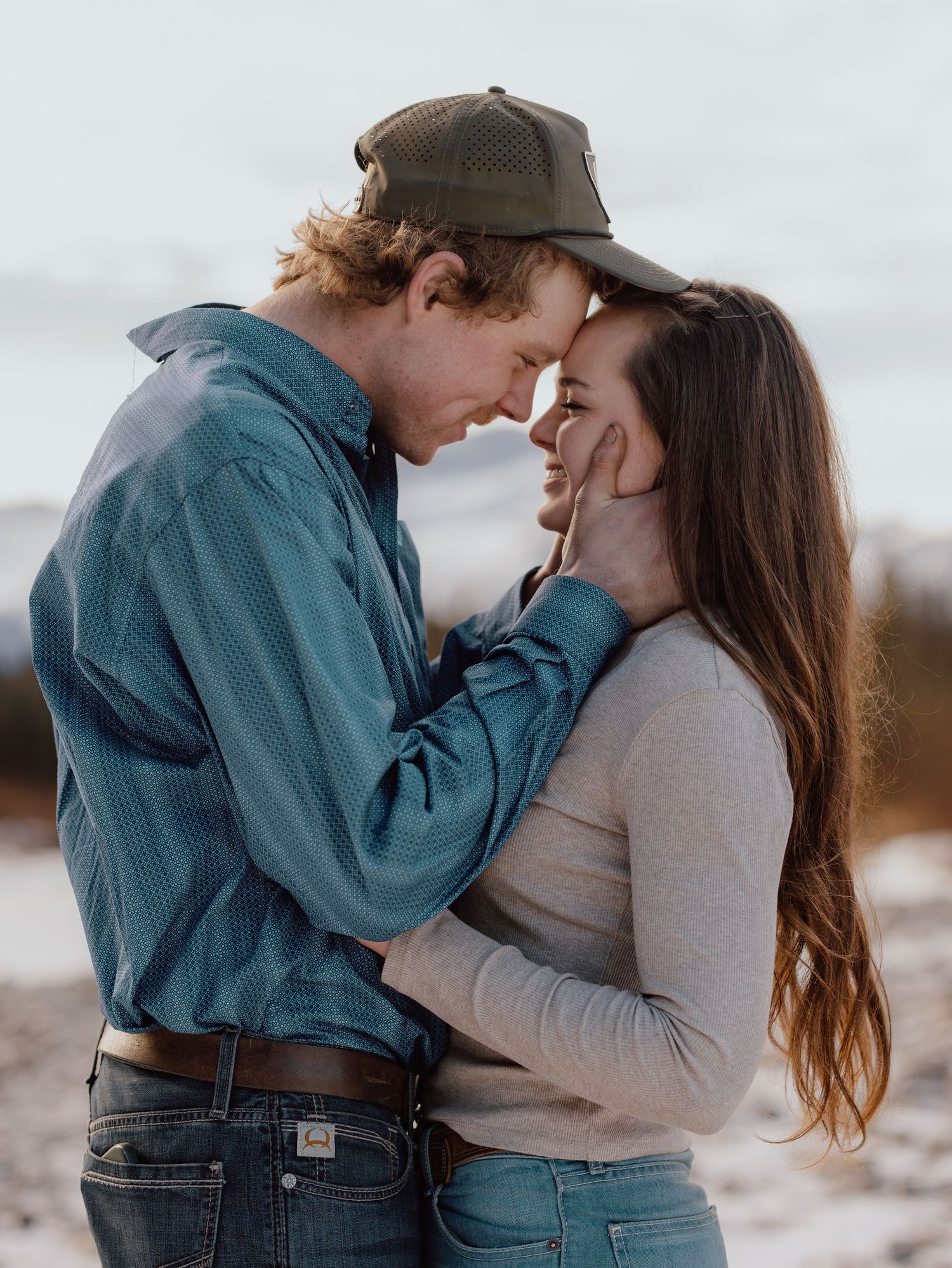Romantic couple sharing an intimate moment outdoors in a snowy landscape, man in teal shirt and cap holding woman's face tenderly.