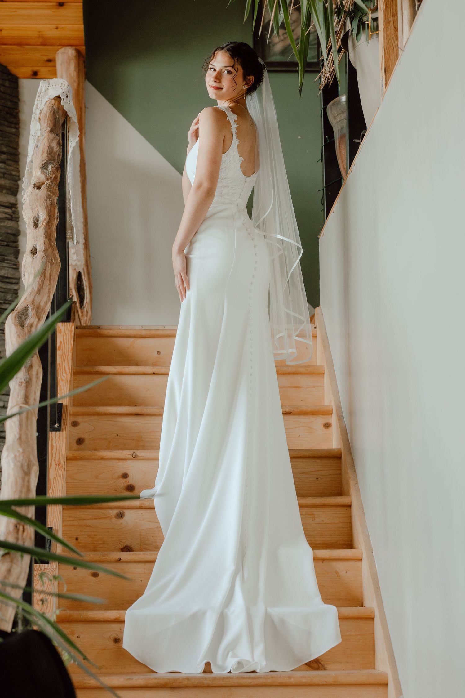 Elegant bride in fitted white gown with button-back detail and veil standing on wooden staircase looking over shoulder.
