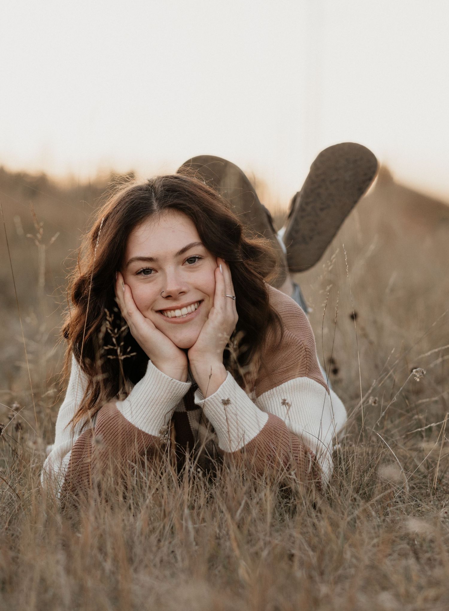 Happy young woman lying in golden grass field, resting face in hands, wearing striped sweater at sunset.