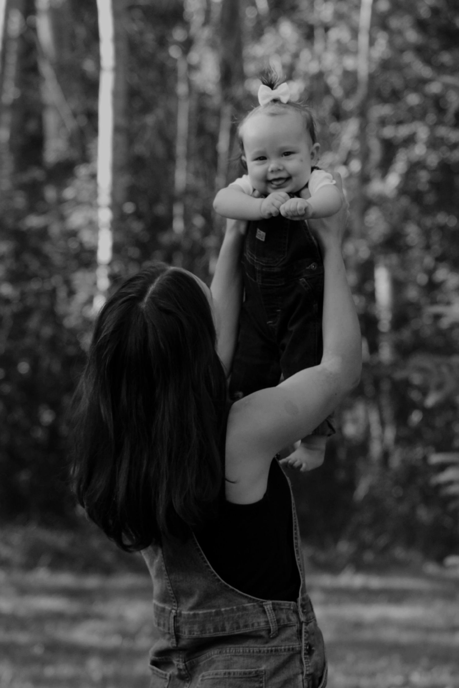 Black and white photo of mother lifting smiling baby girl with bow in a birch tree forest.