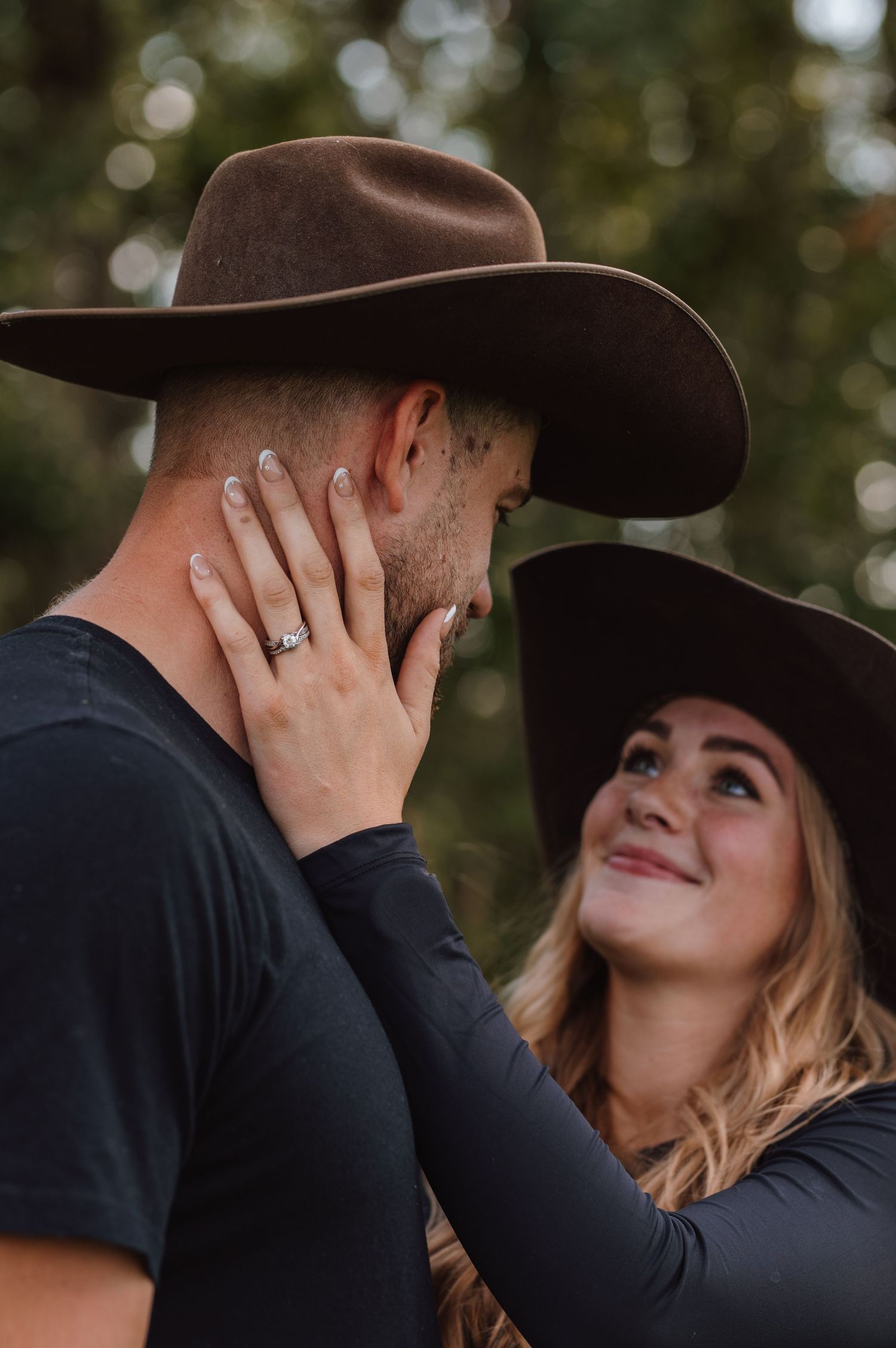 Couple in matching black cowboy hats sharing a romantic moment outdoors, woman wearing engagement ring touching mans face.