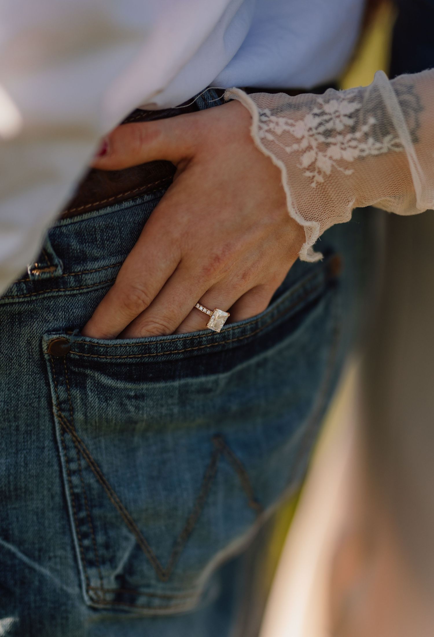 Close-up of a woman's hand in jeans pocket wearing a diamond engagement ring with lace sleeve detail.