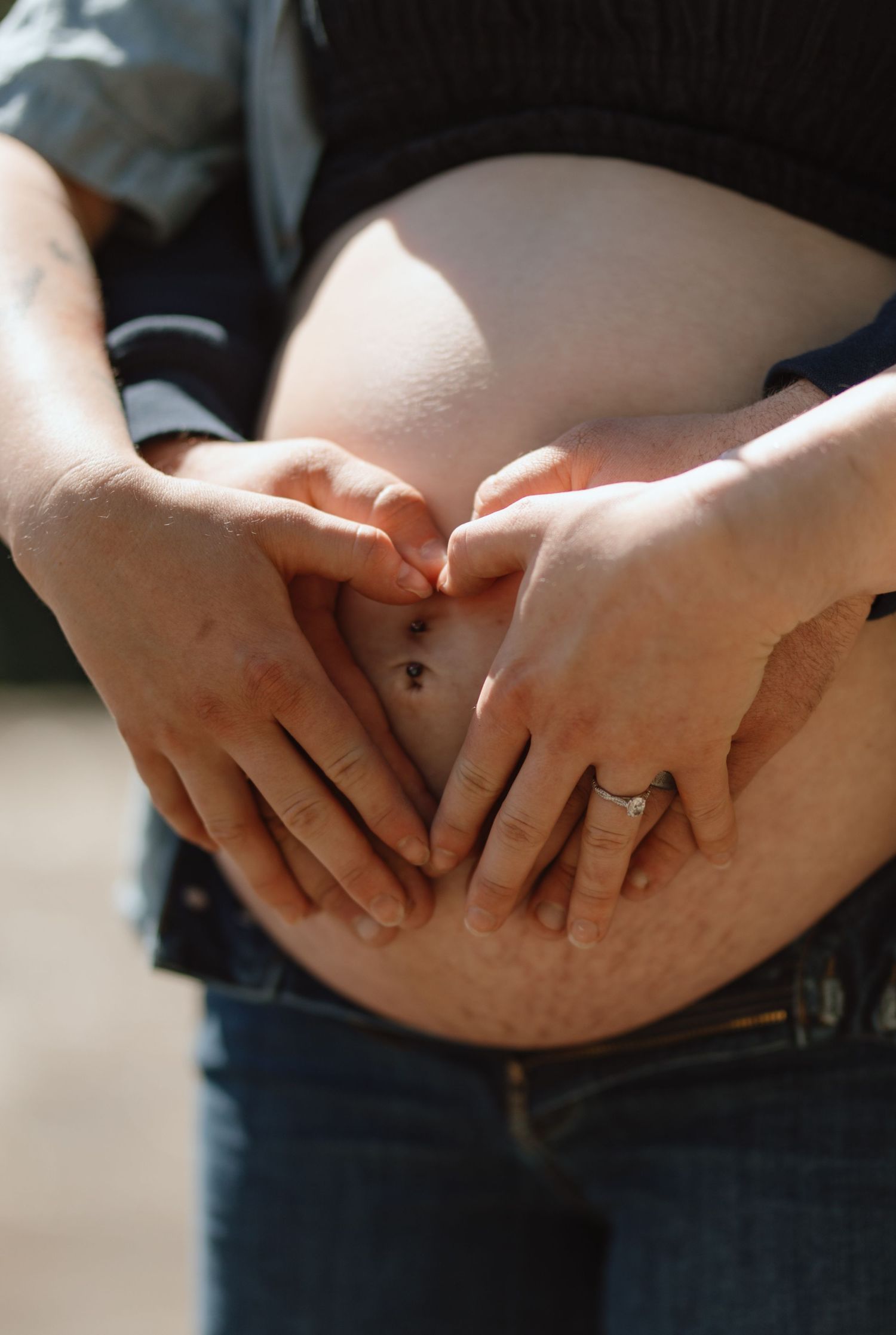 Couple forming heart shape with hands on pregnant belly, woman wearing engagement ring outdoors.