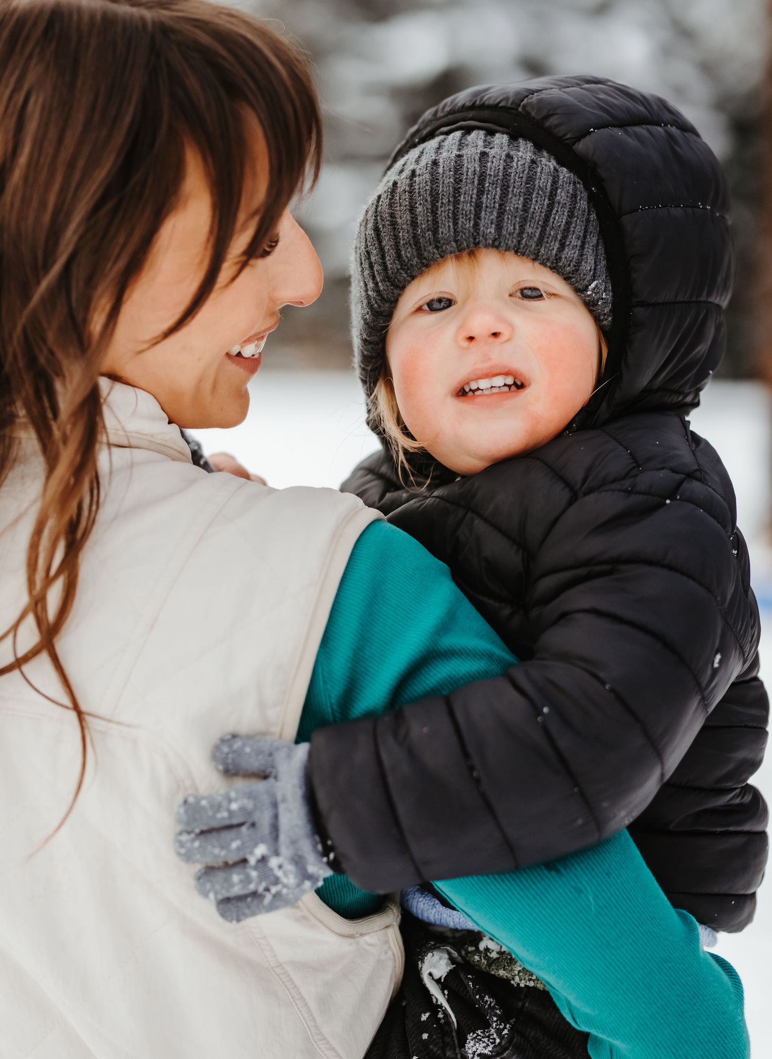 Mother holding bundled toddler in black puffer jacket and grey knit hat in winter snow.