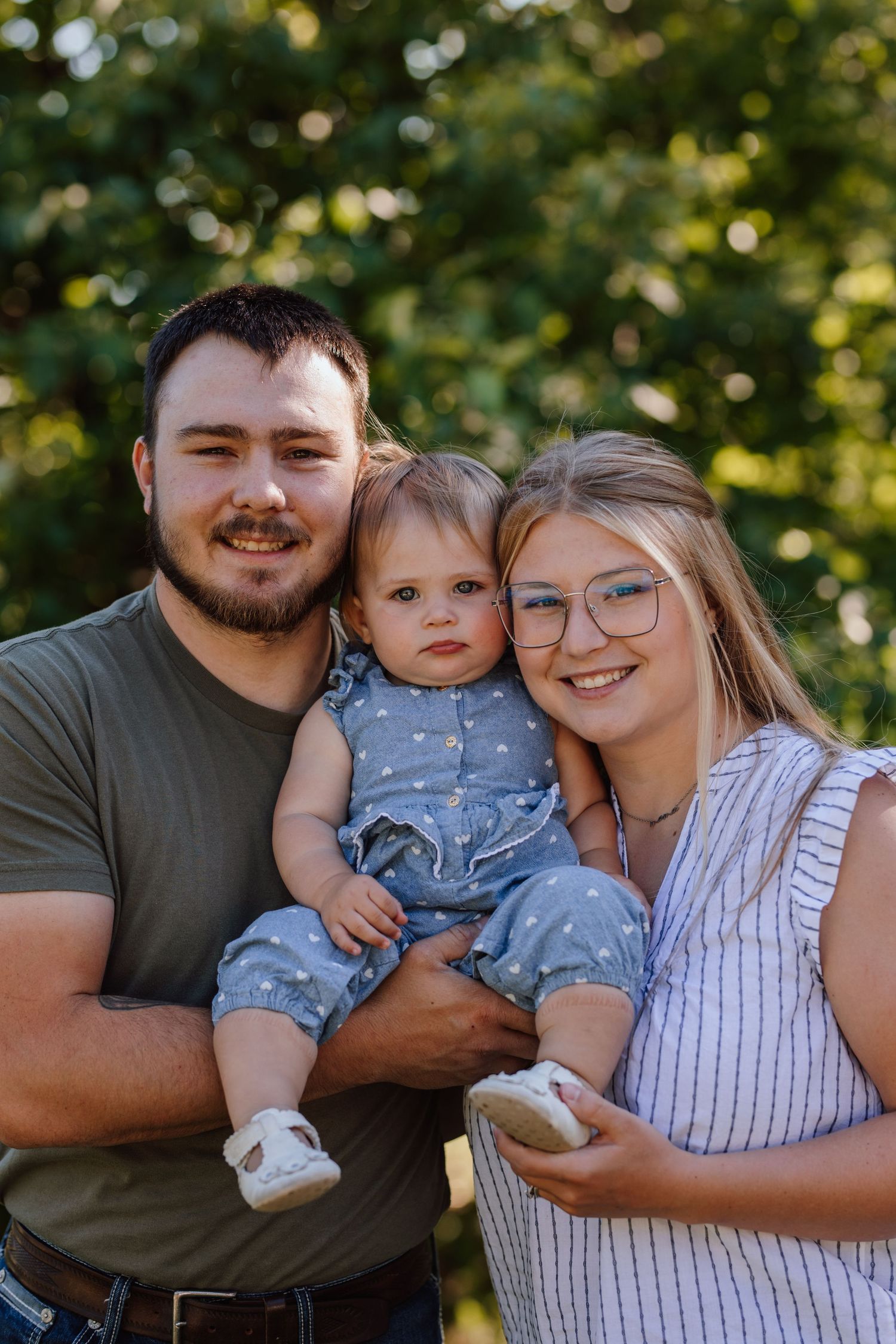Young family portrait outdoors, parents smiling and holding baby in blue romper with green trees background.