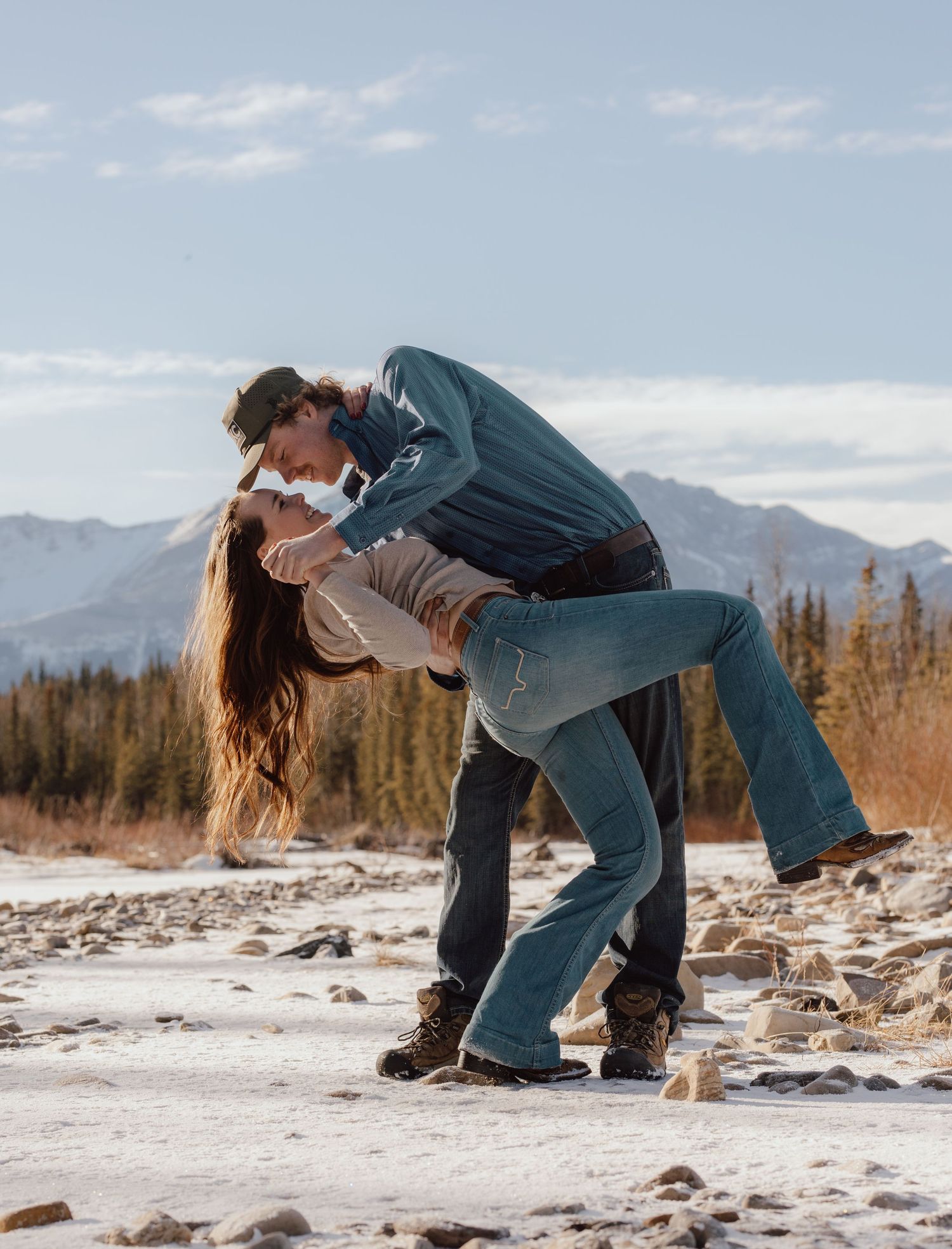 Couple shares a passionate dip kiss in a snowy mountain landscape with trees in the background.