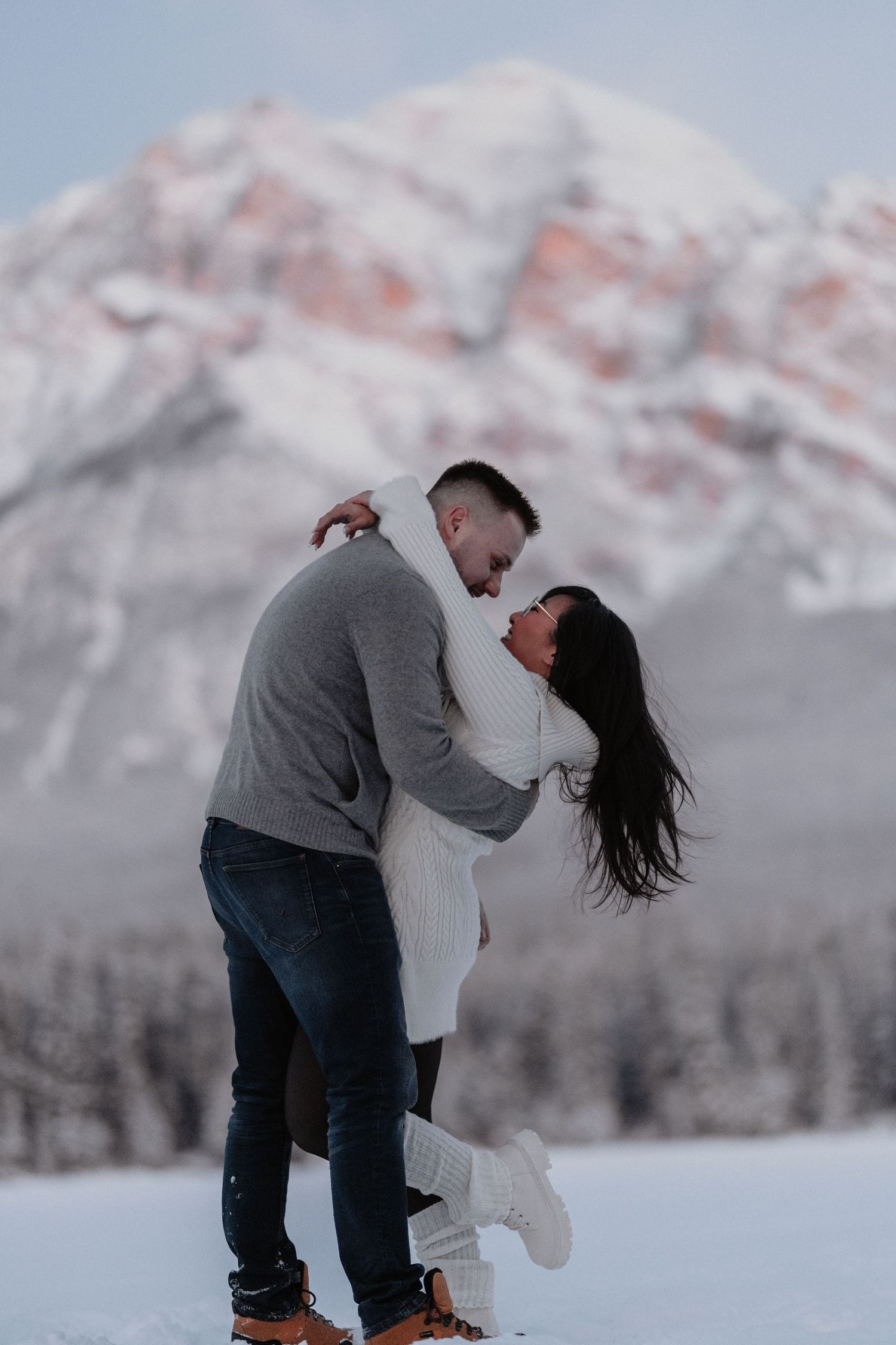 Couple embracing and laughing in snow with dramatic mountain peak backdrop in winter.