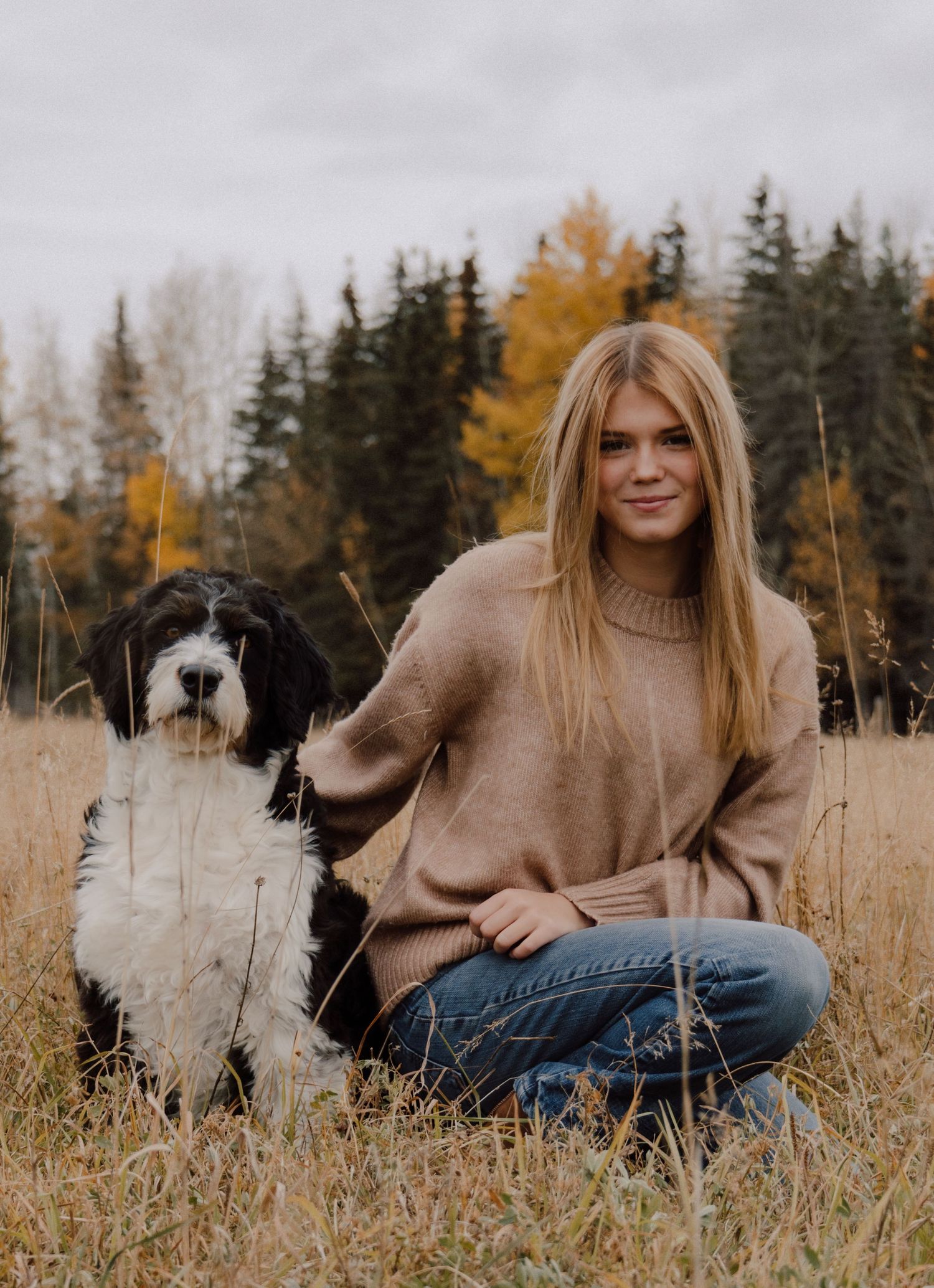 Young woman in beige sweater sitting in autumn field with black and white dog, surrounded by fall foliage.