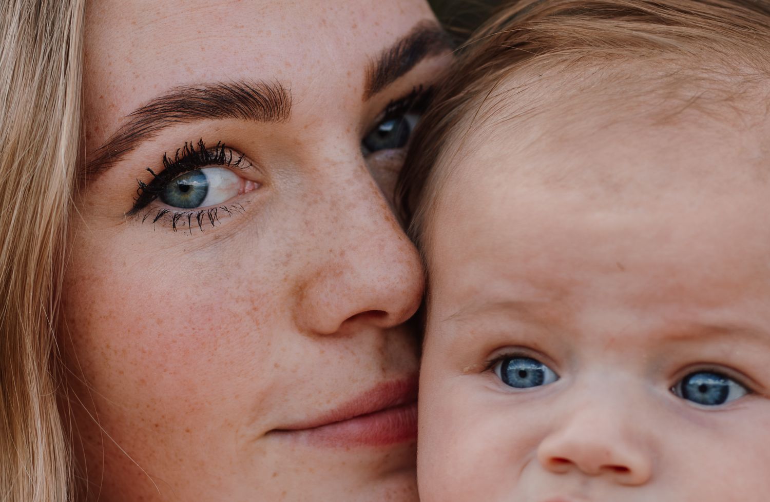 Close-up portrait of a young woman with blue eyes and freckles cheek-to-cheek with a blue-eyed baby.