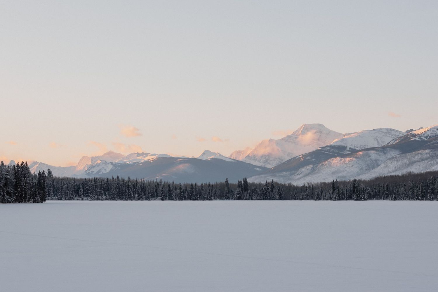 Snow-covered frozen lake with evergreen forest and pink-hued mountain peaks at dusk in winter wilderness.