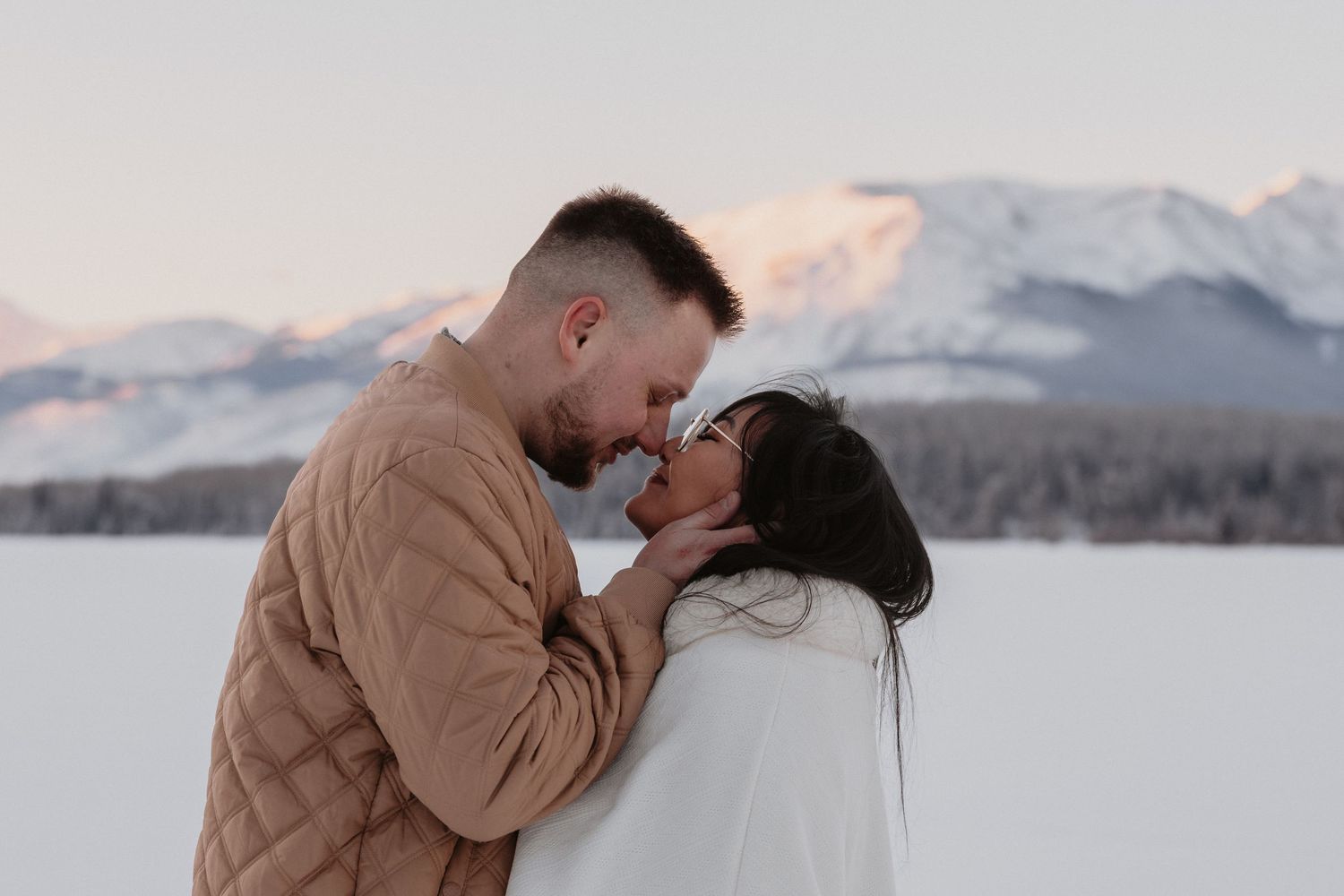 Couple sharing a romantic moment in snowy mountain landscape, wearing cozy winter sweaters at sunset.