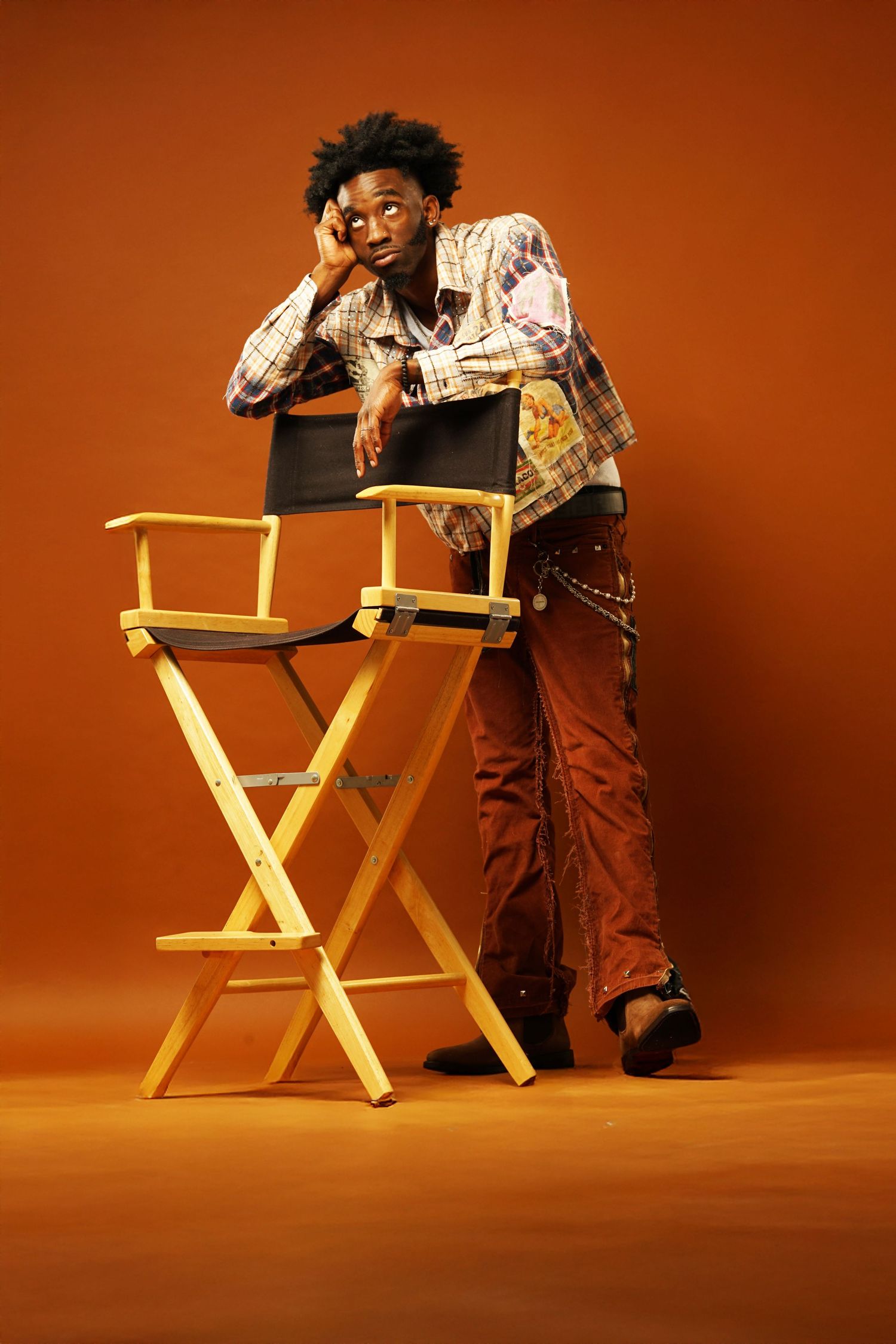 Casual pose leaning on yellow director's chair wearing patchwork jacket and orange pants.