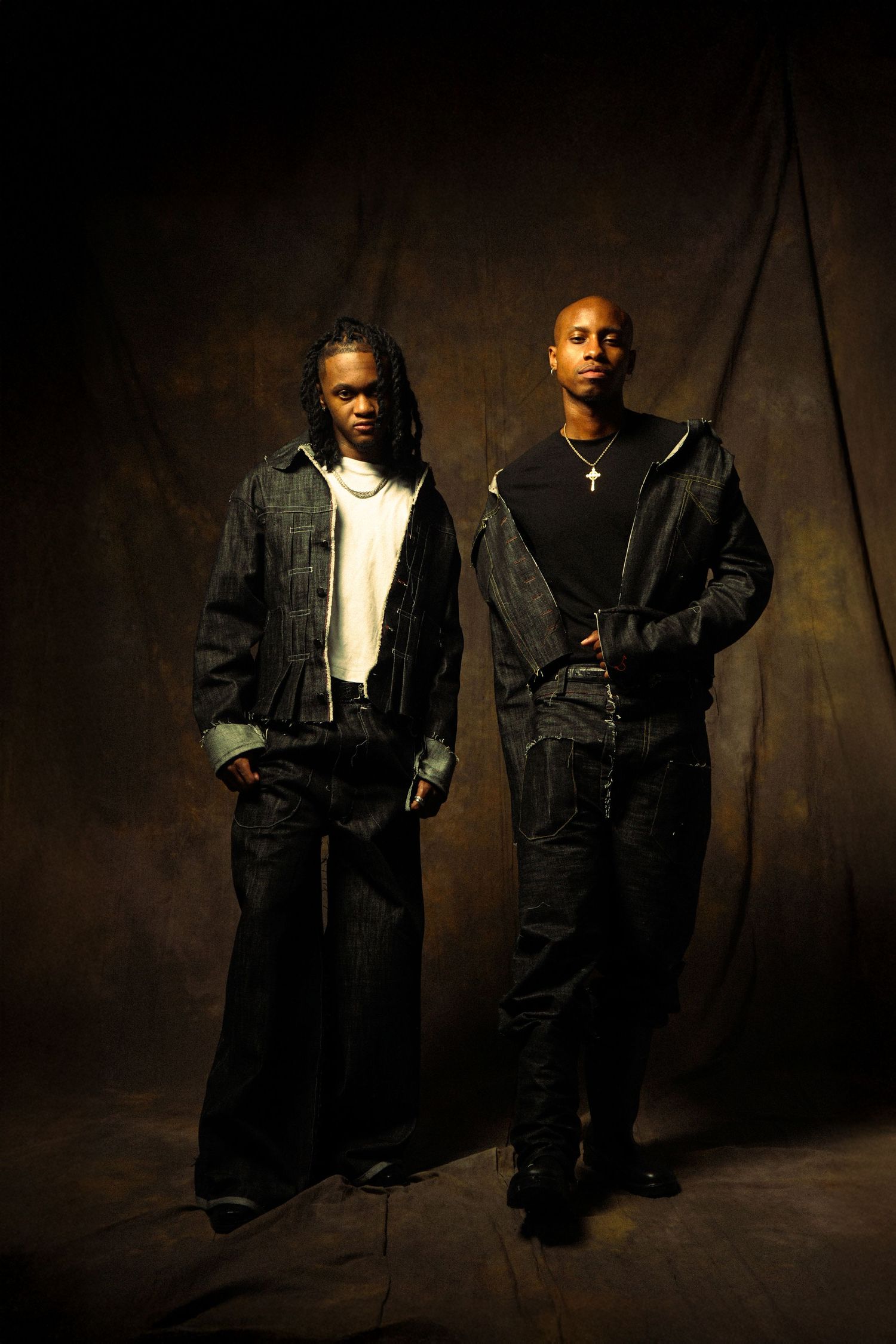 Two men pose against dark background wearing denim jackets and casual attire in moody portrait lighting.