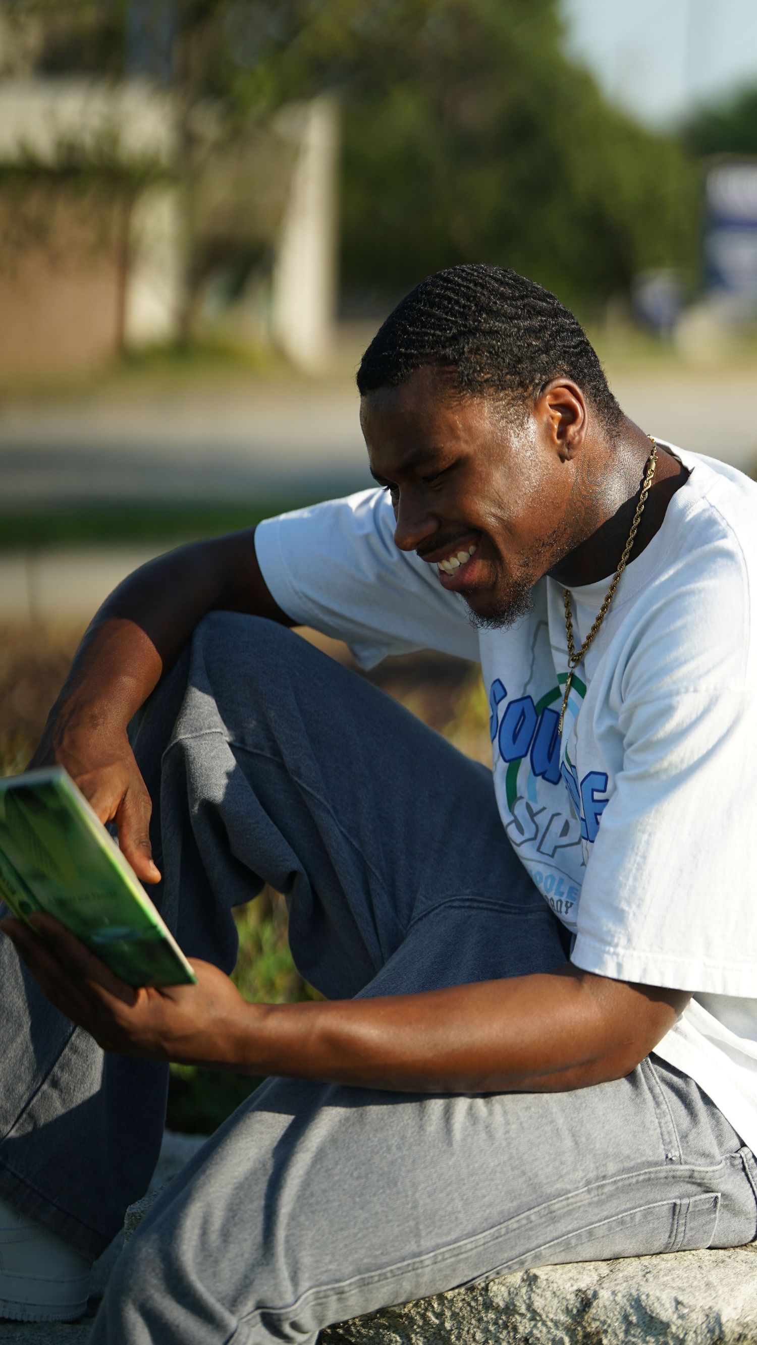 Person in white t-shirt sits outdoors reading a book with engaged expression.