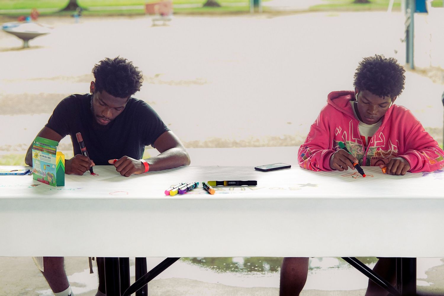 Two people sit at a white folding table outdoors, drawing with markers on paper under a shaded canopy.