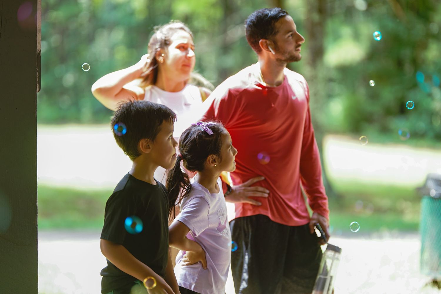 Family of four enjoying a sunny day outdoors with bubbles floating in the air at a lush green park.