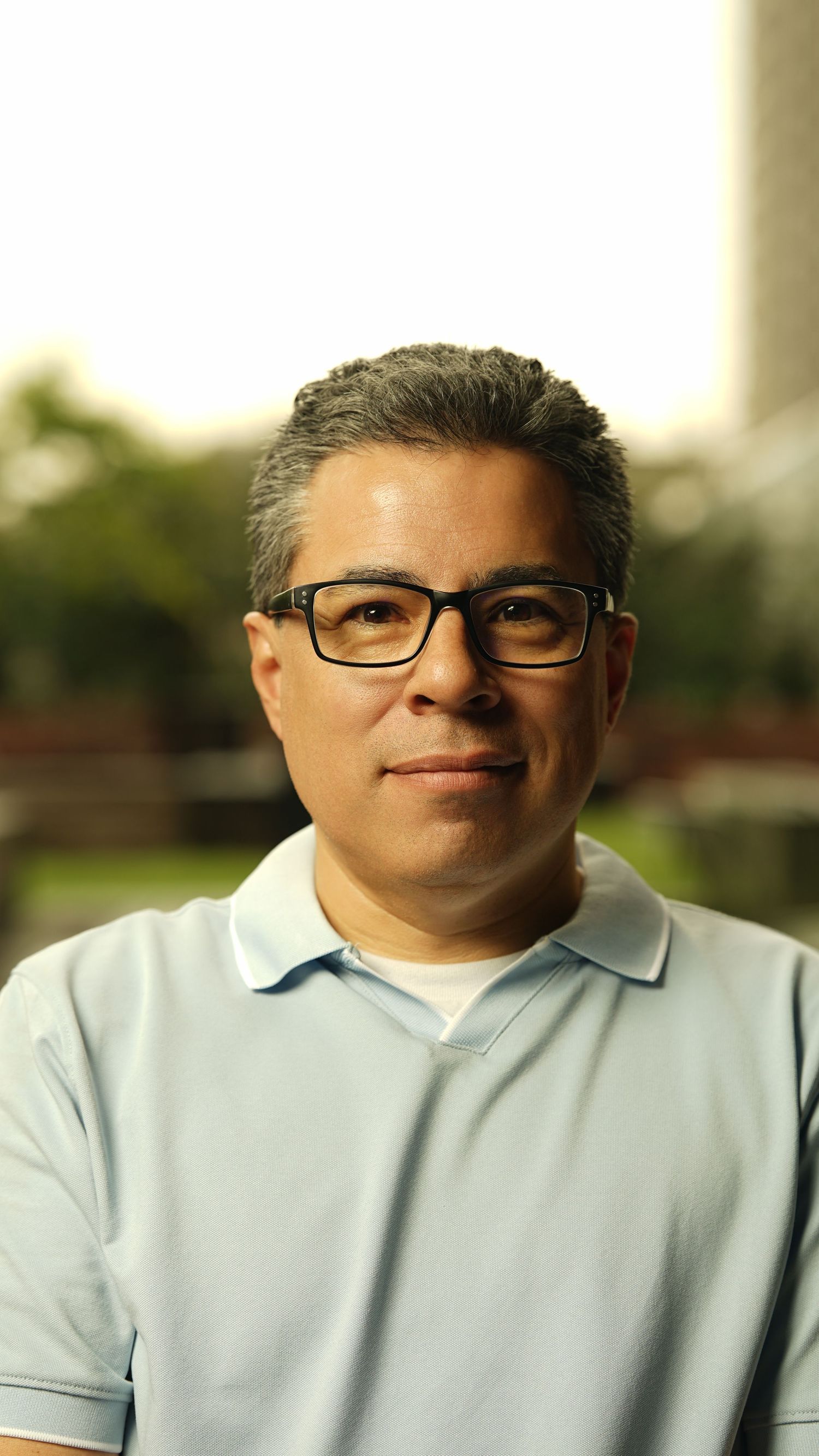 Person in light blue polo shirt and glasses smiling at camera during outdoor portrait session.