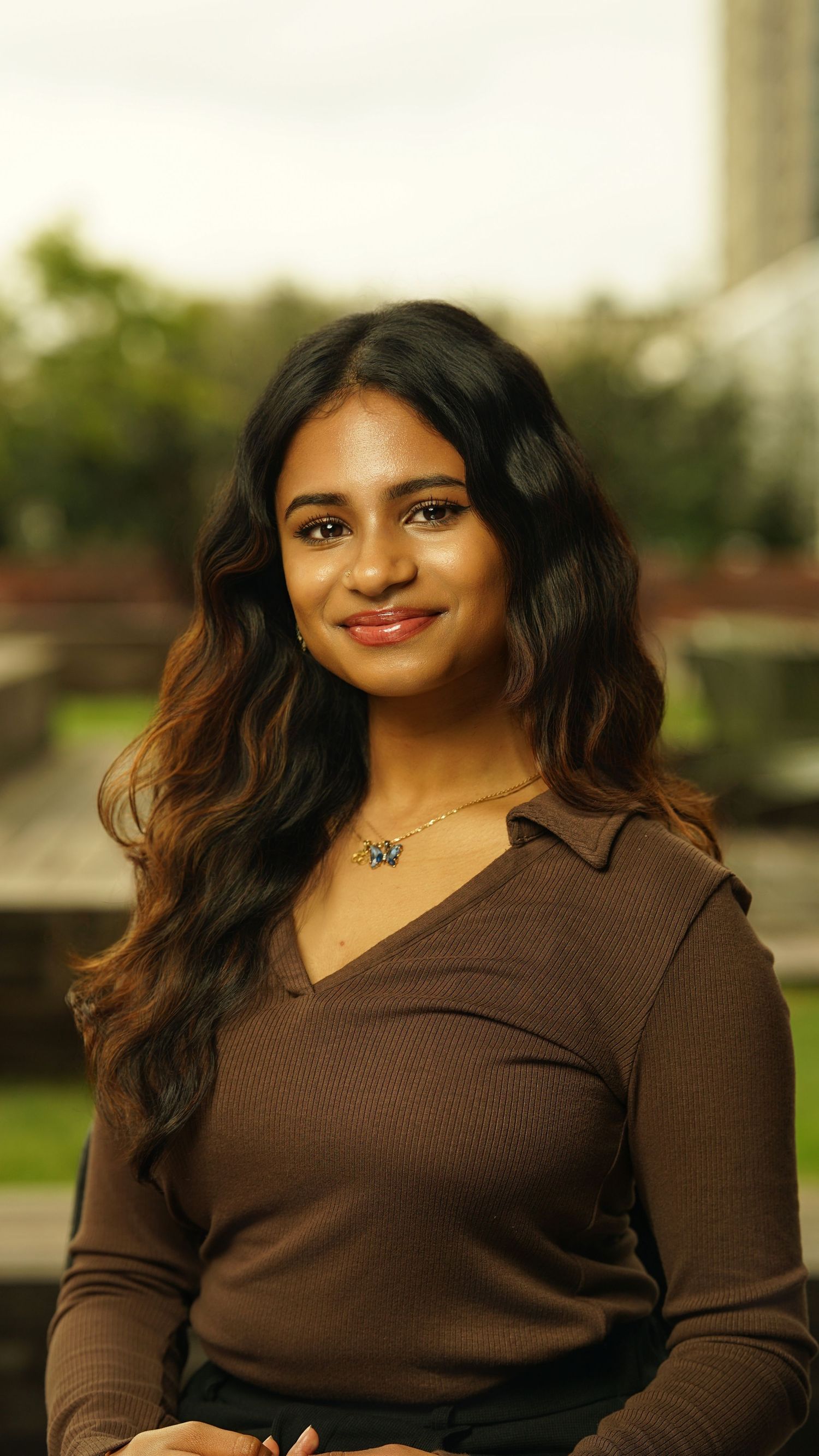 Professional portrait of someone in a brown sweater smiling warmly against a blurred outdoor background.