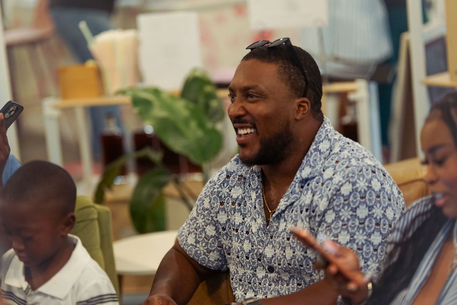 Smiling Black man in a blue patterned shirt laughing during a casual indoor gathering or event.