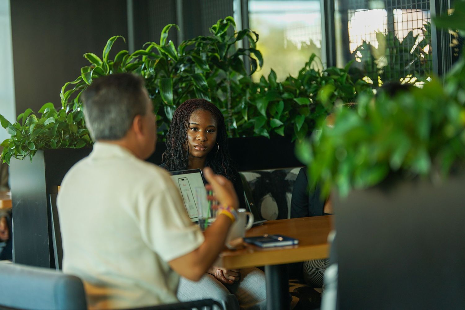 A Black woman and an older man have a focused one-on-one meeting at a table with plants.