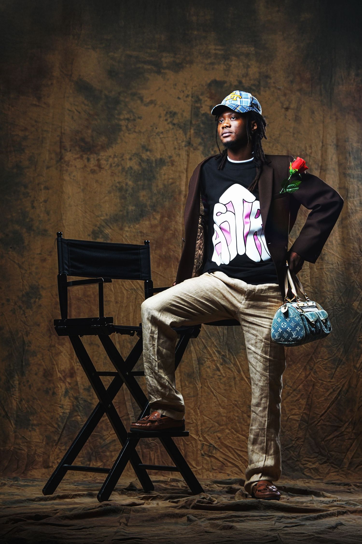 Model seated in director's chair wearing casual attire against rustic brown background.