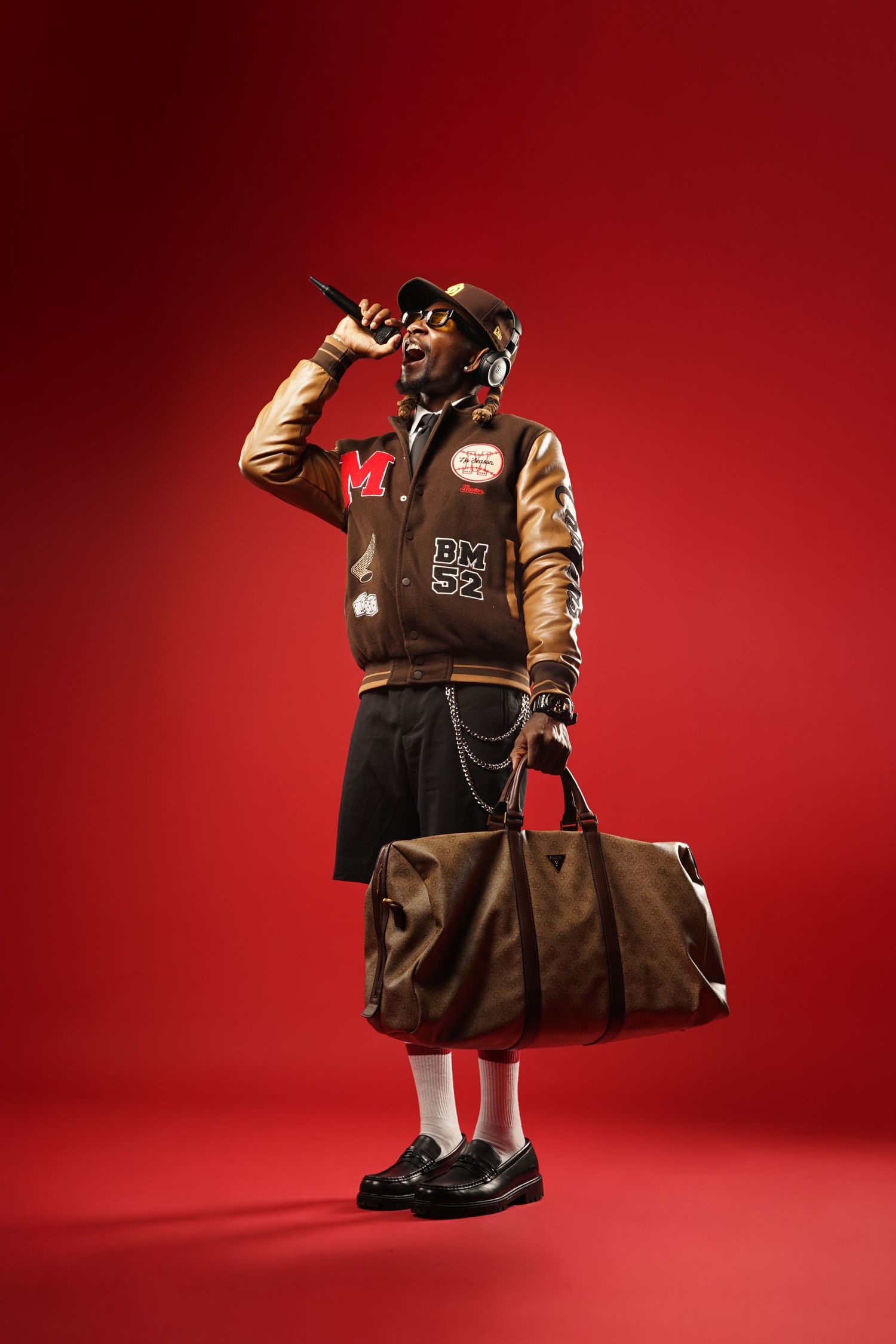 Stylish varsity jacket outfit with brown duffel bag against red backdrop in vintage prep school fashion aesthetic.