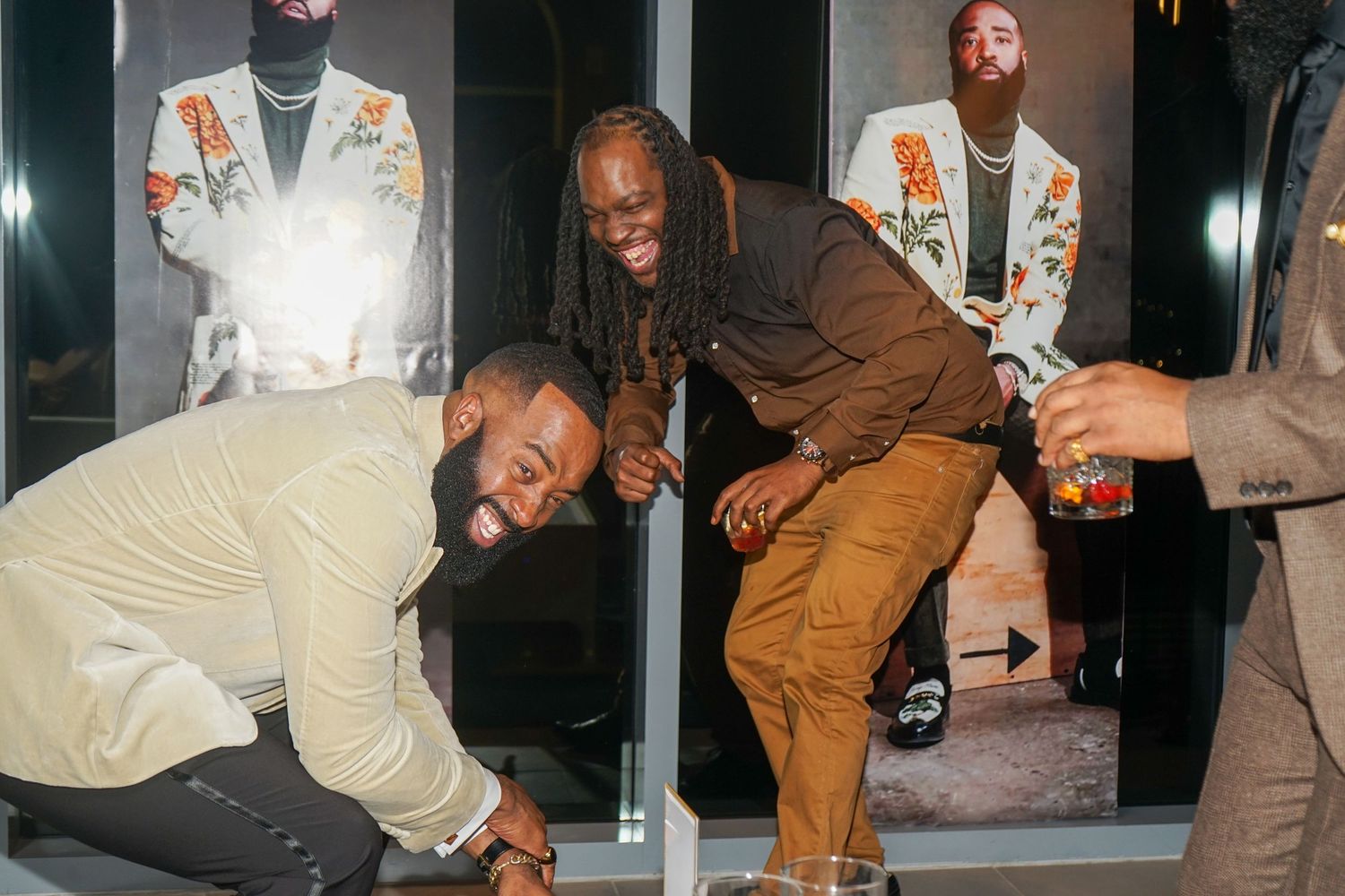 Three stylish men laughing and posing at an indoor event, one bending down, wearing fashionable outfits.