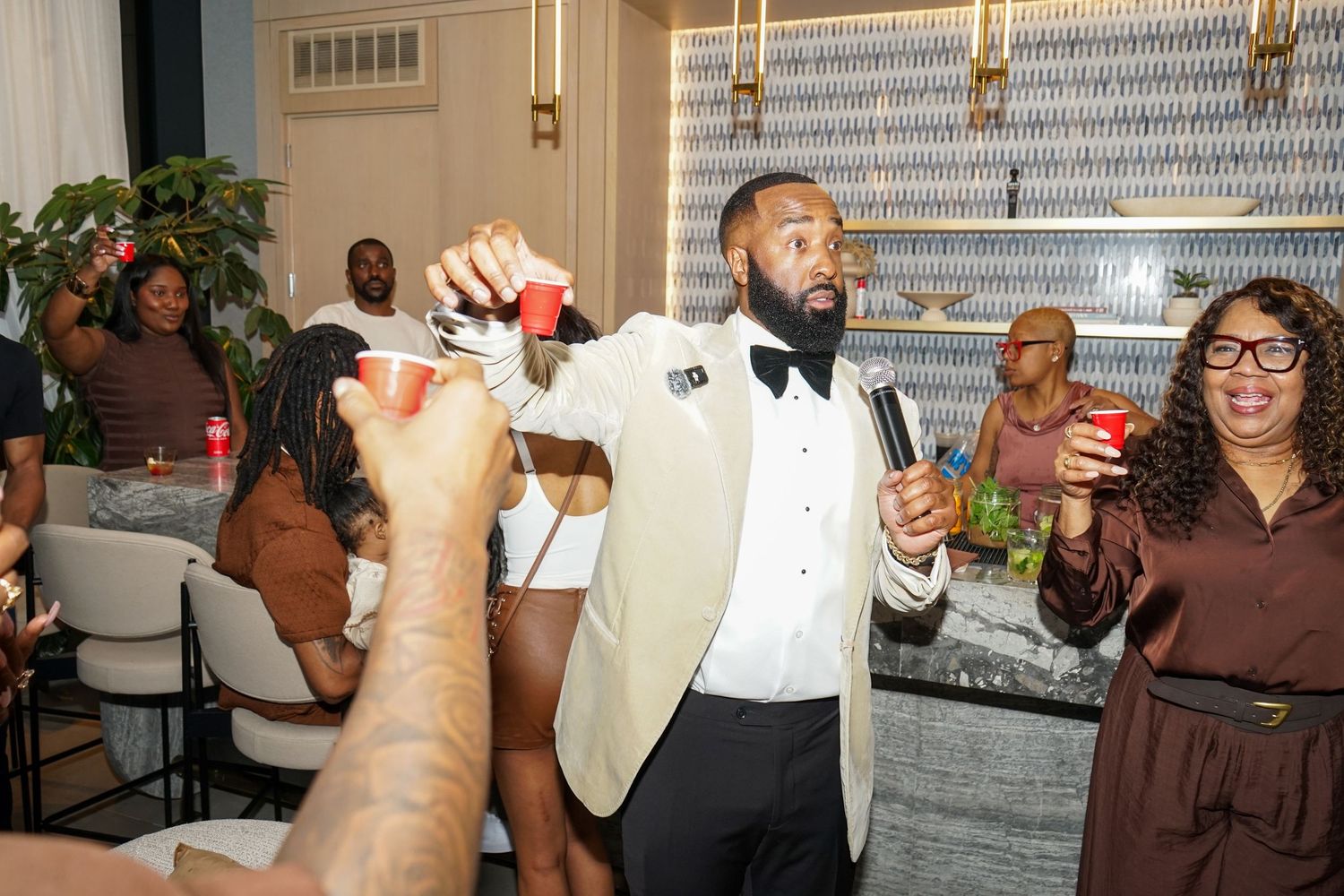 Man in white tuxedo jacket toasting with red cups at a lively indoor party celebration.