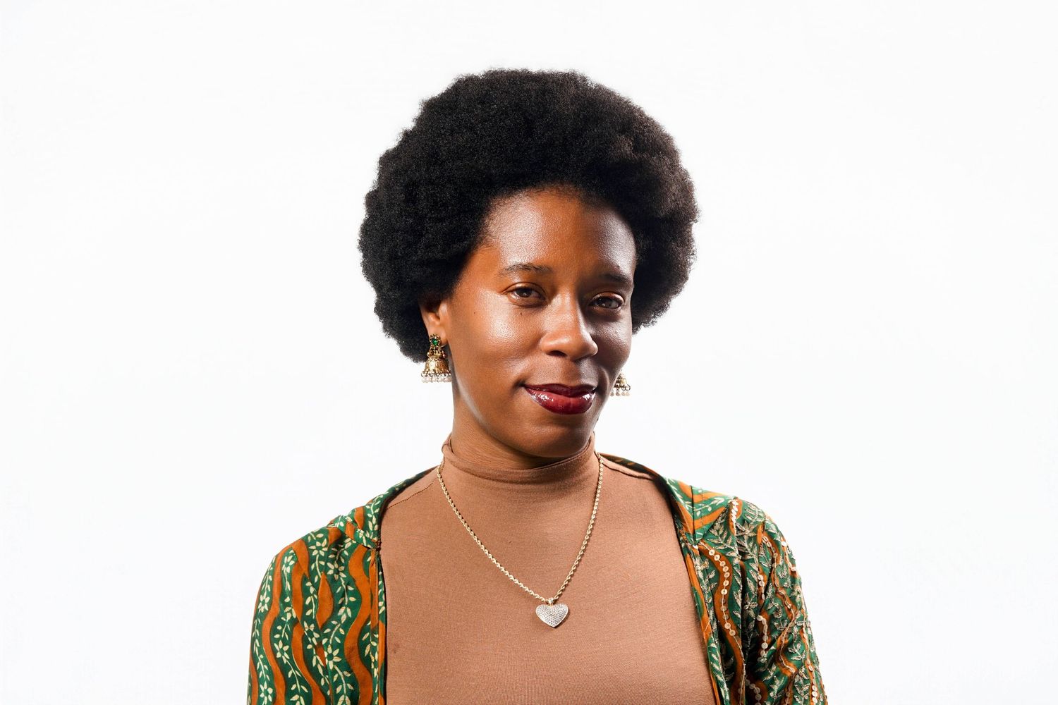 Young Black woman with afro hair wearing a patterned green jacket and heart necklace, smiling against white background.