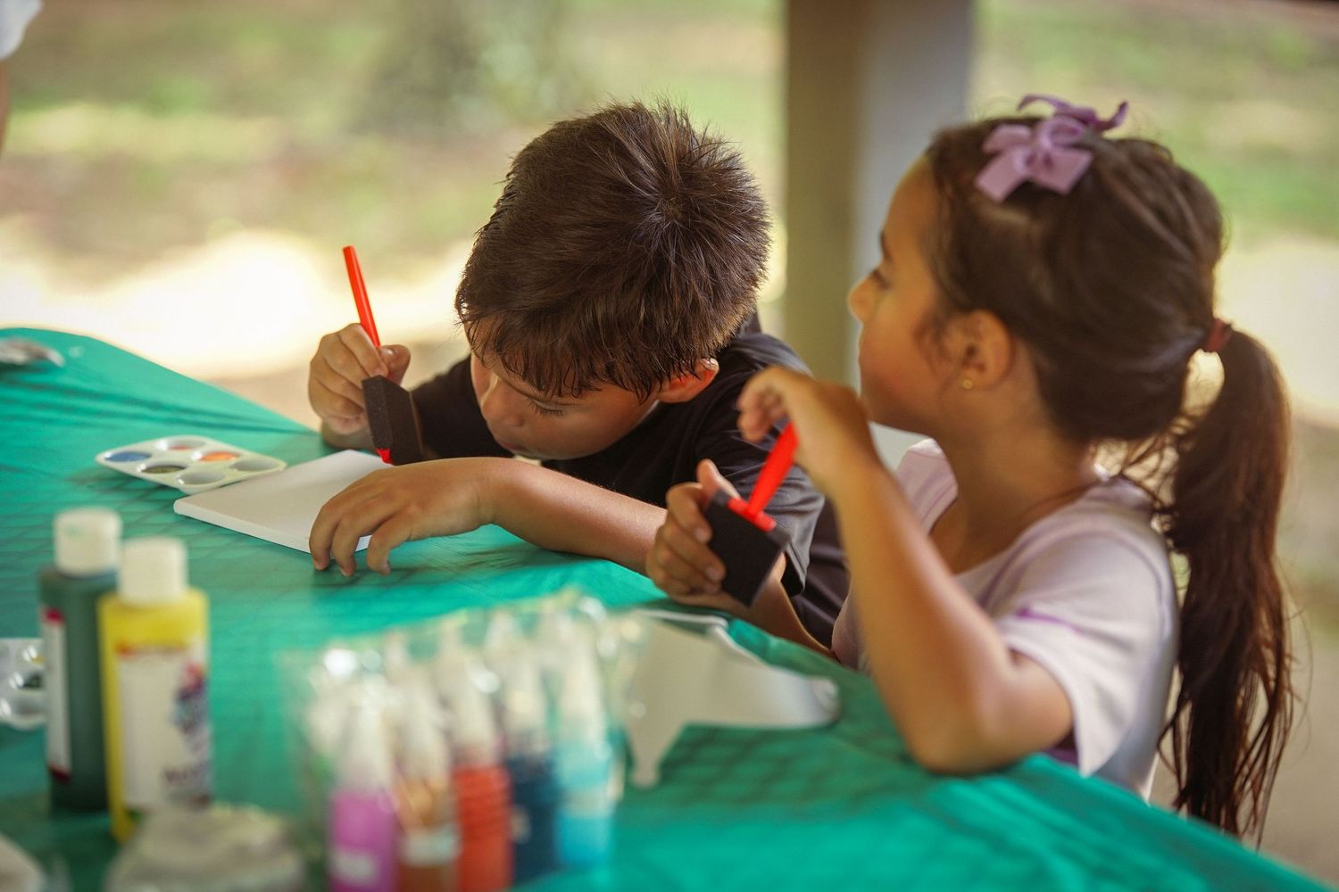 Two children painting and drawing together at a table covered with a green tablecloth and colorful paint bottles.