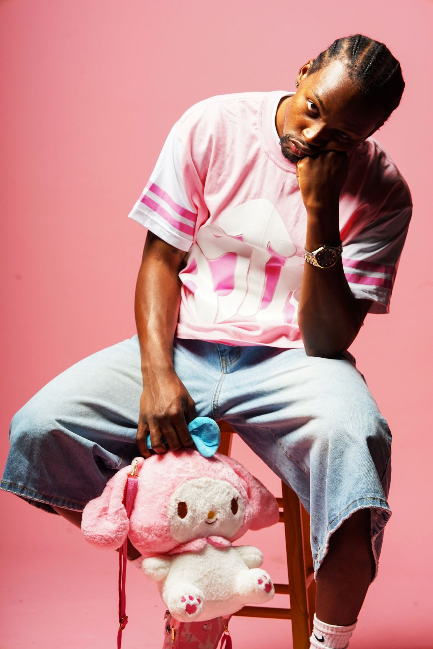 Person sitting with pink plush toy on chair against pink background wearing casual outfit.
