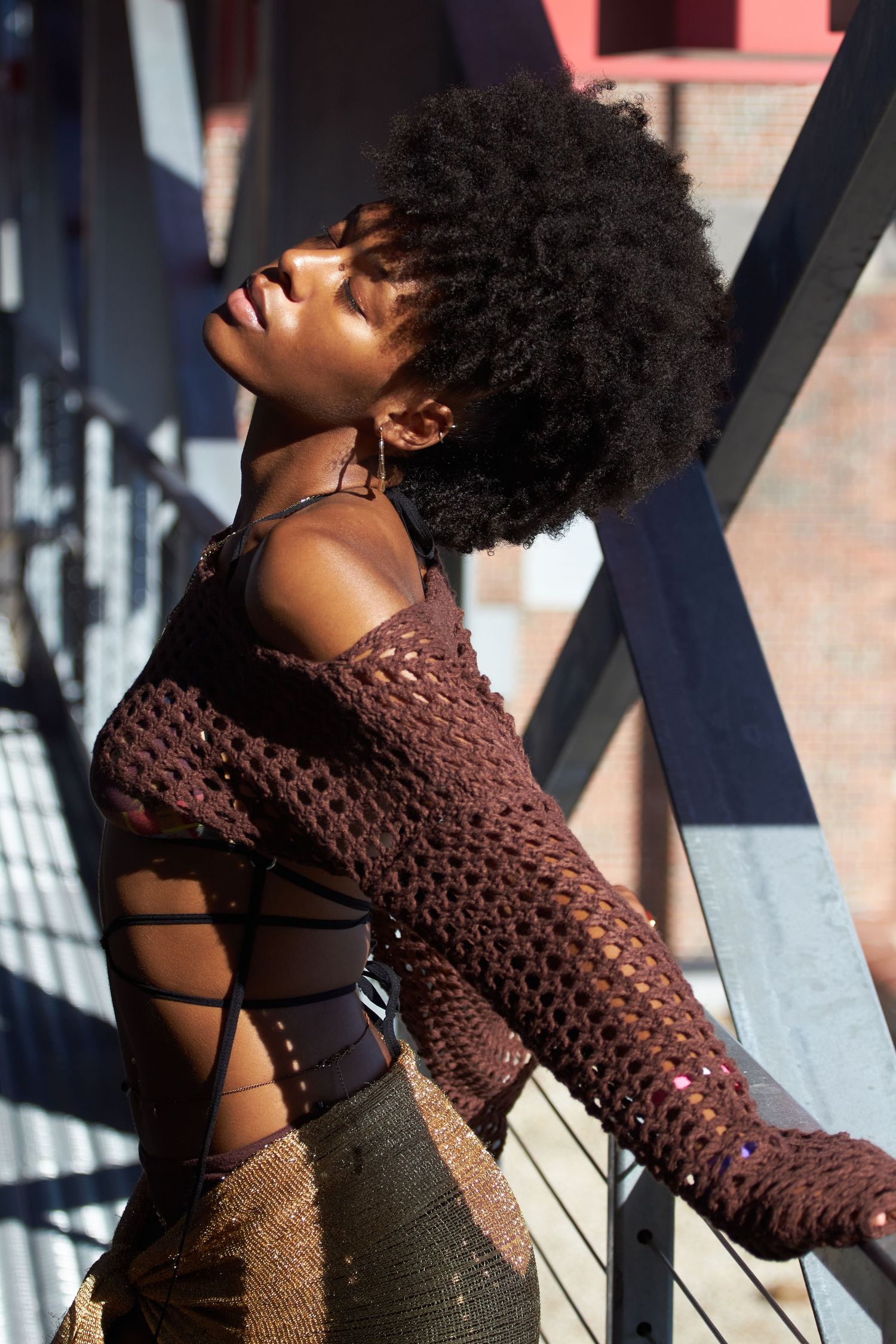 Person wearing brown sweater and natural hairstyle poses against architectural backdrop with dramatic lighting.