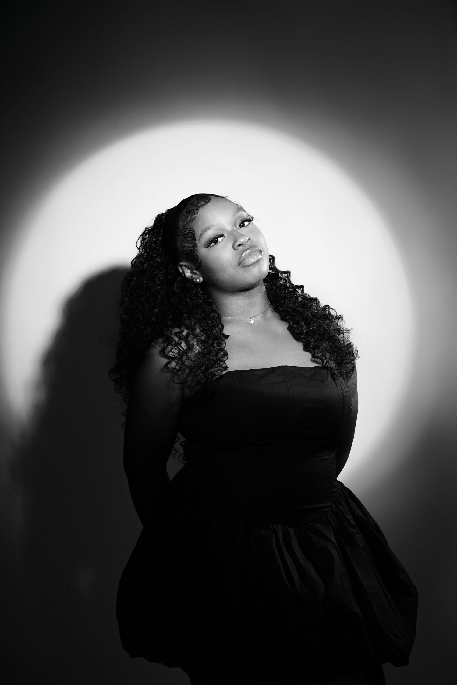 Black and white portrait of a young woman in a black dress posing confidently against a spotlight background.