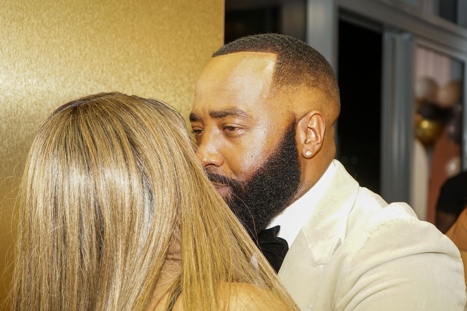 A man in a white tuxedo with a beard shares an intimate moment with a blonde woman at a formal event.