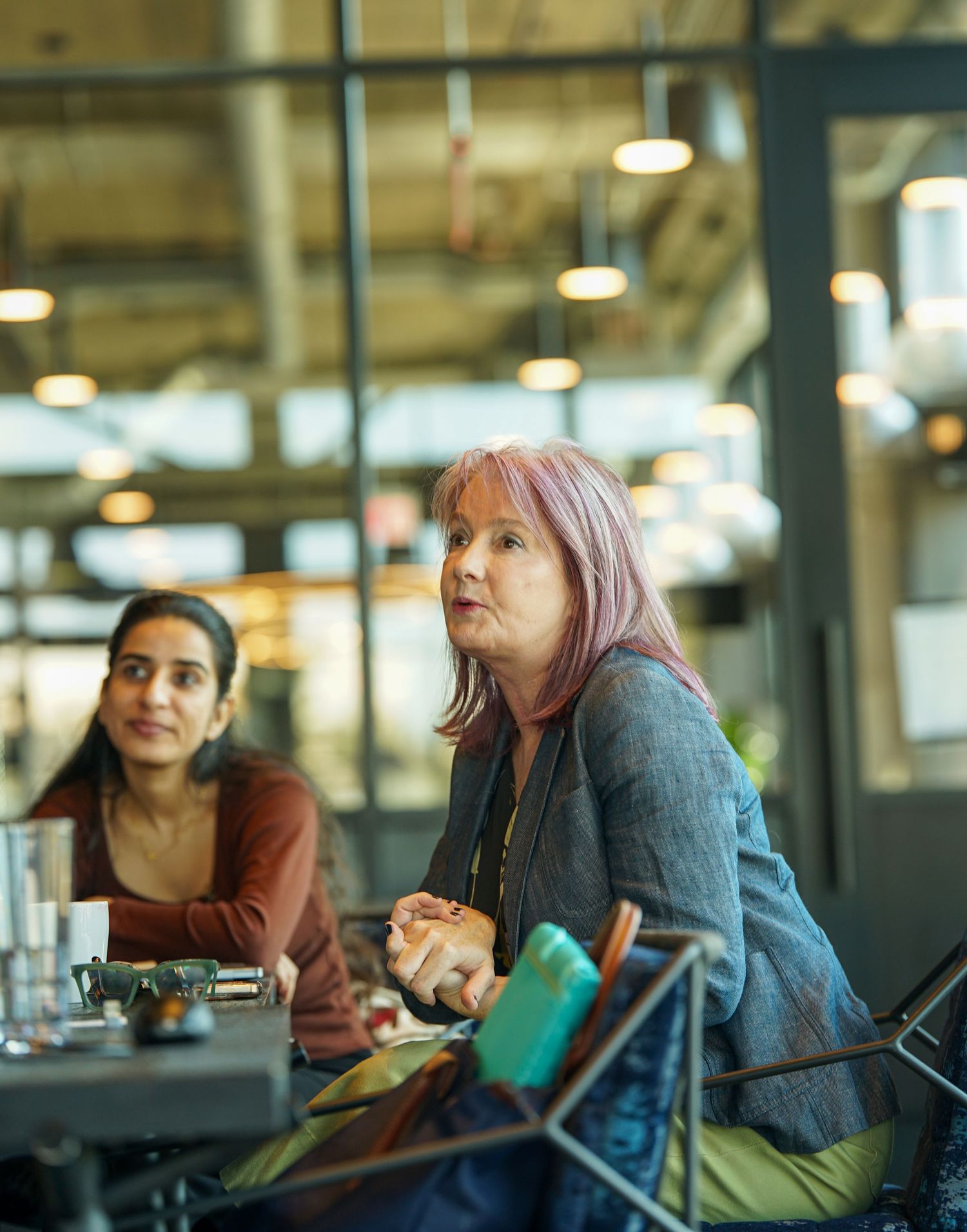 Two women engage in discussion at a modern cafe, one with pink hair in a gray blazer.