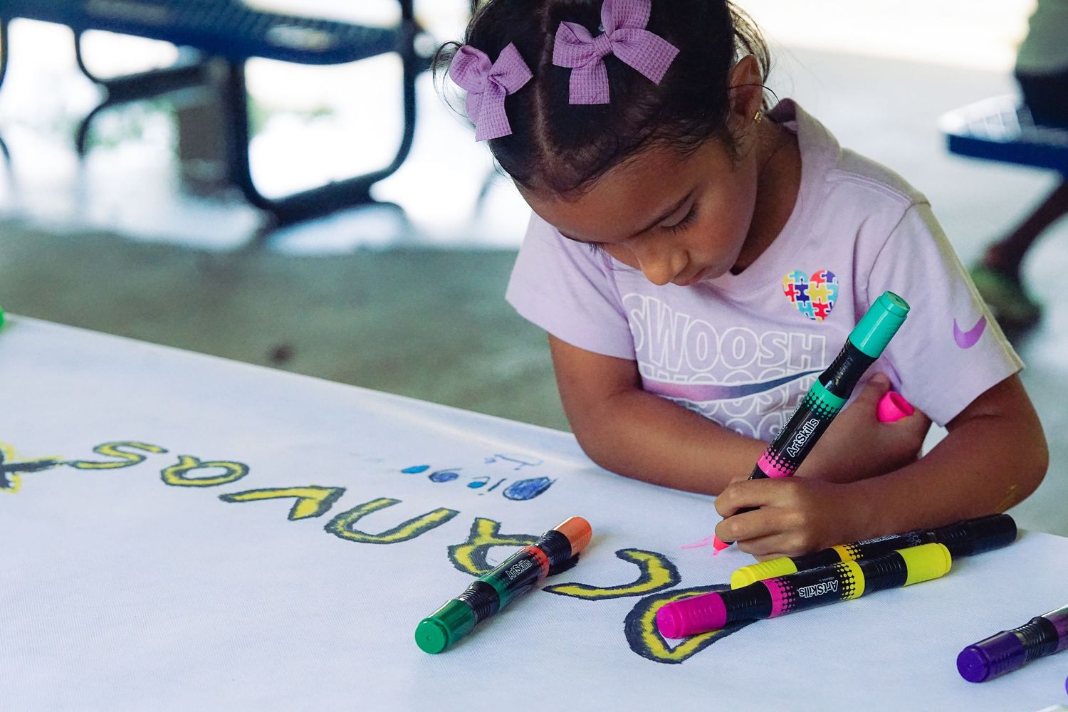 Young girl with pink bows colors on large paper banner with bright markers at outdoor event.