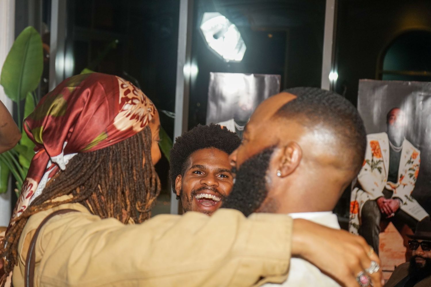 Three people share a warm embrace at a social gathering, smiling joyfully in a dimly lit venue.