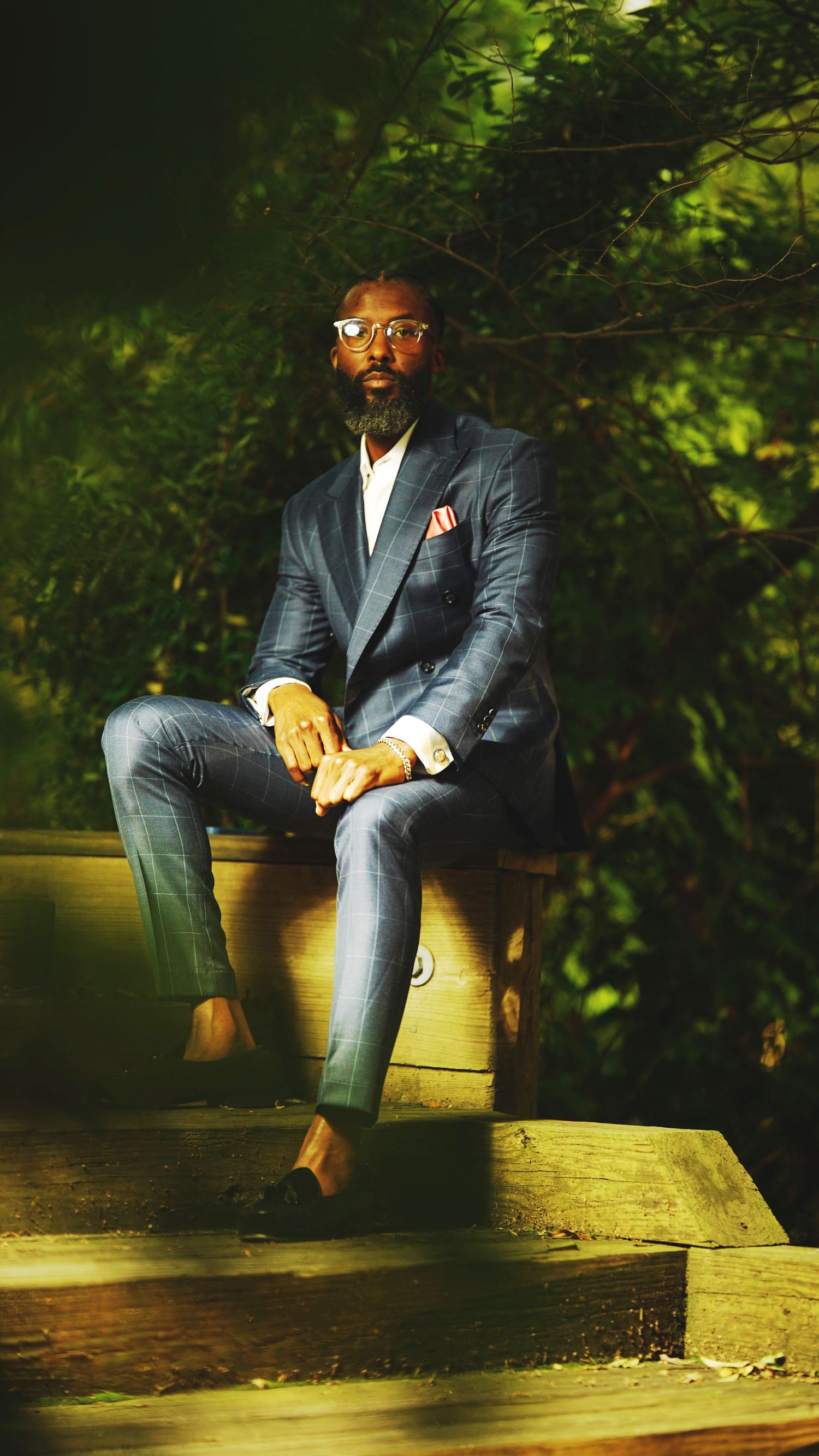 Stylish figure in a dark suit and glasses sits on a concrete ledge in an outdoor evening setting.