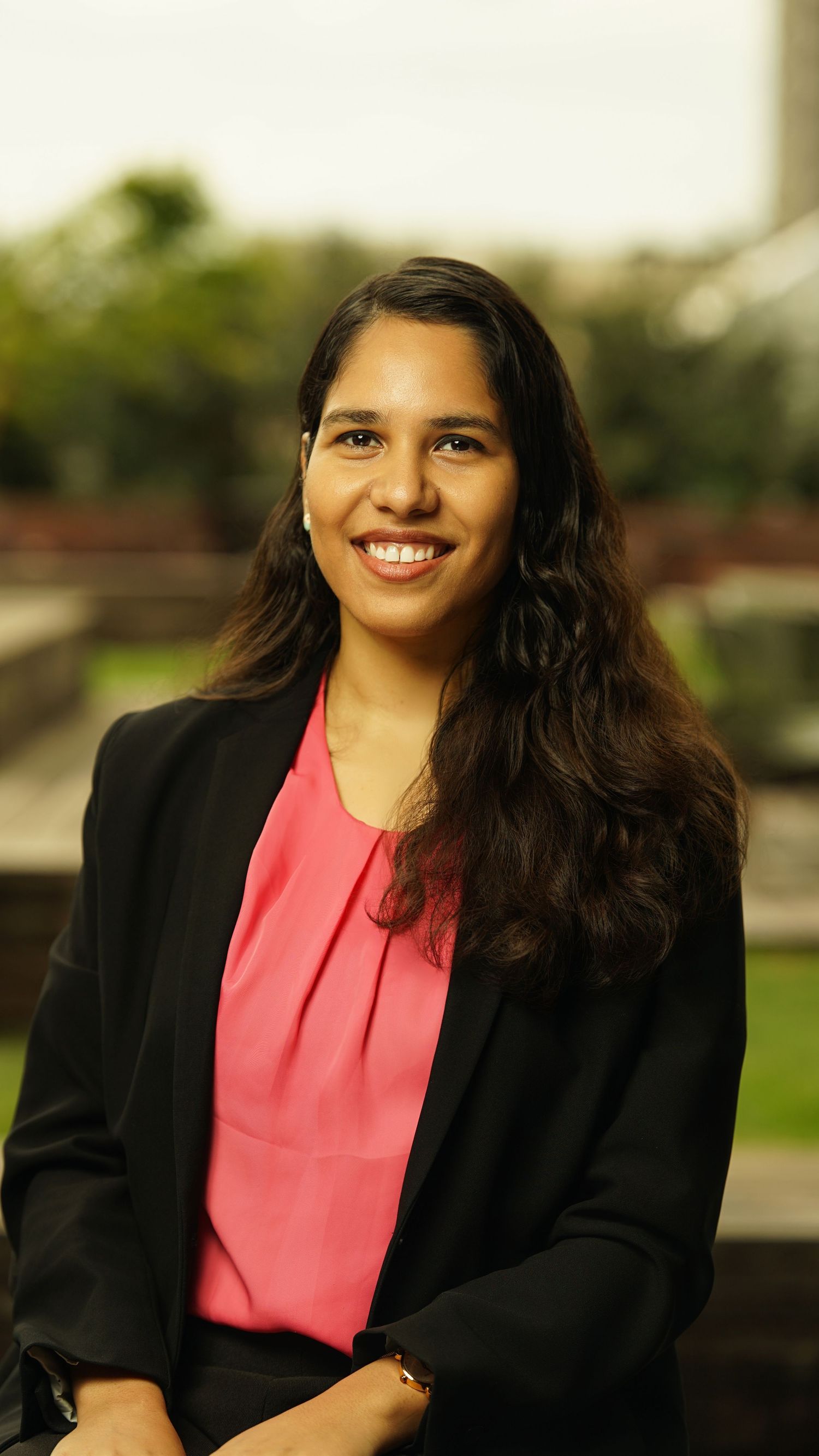 Professional portrait of person wearing pink blouse and black blazer with natural outdoor backdrop.