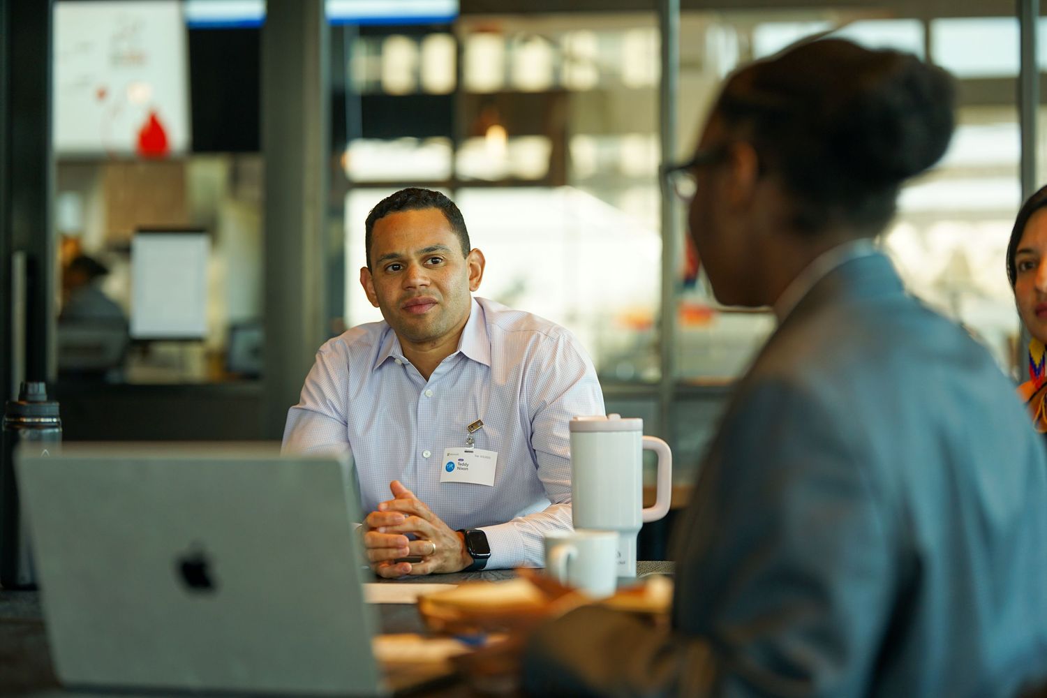 A professional man with a badge listens attentively during a business meeting with a laptop nearby.